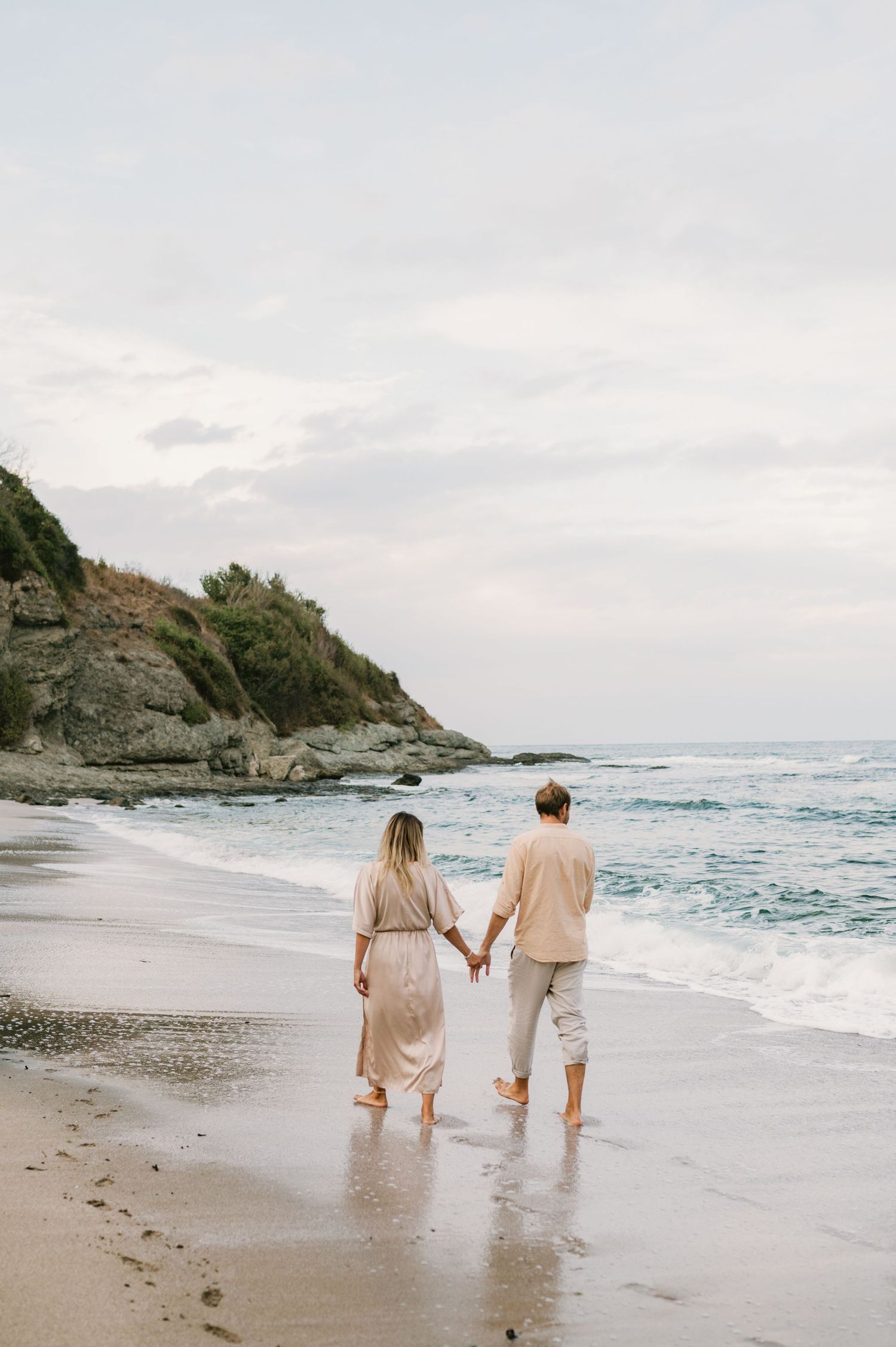 Couple holding hands walking barefoot on a sandy beach at dusk with rocky cliffs and ocean waves in the background.