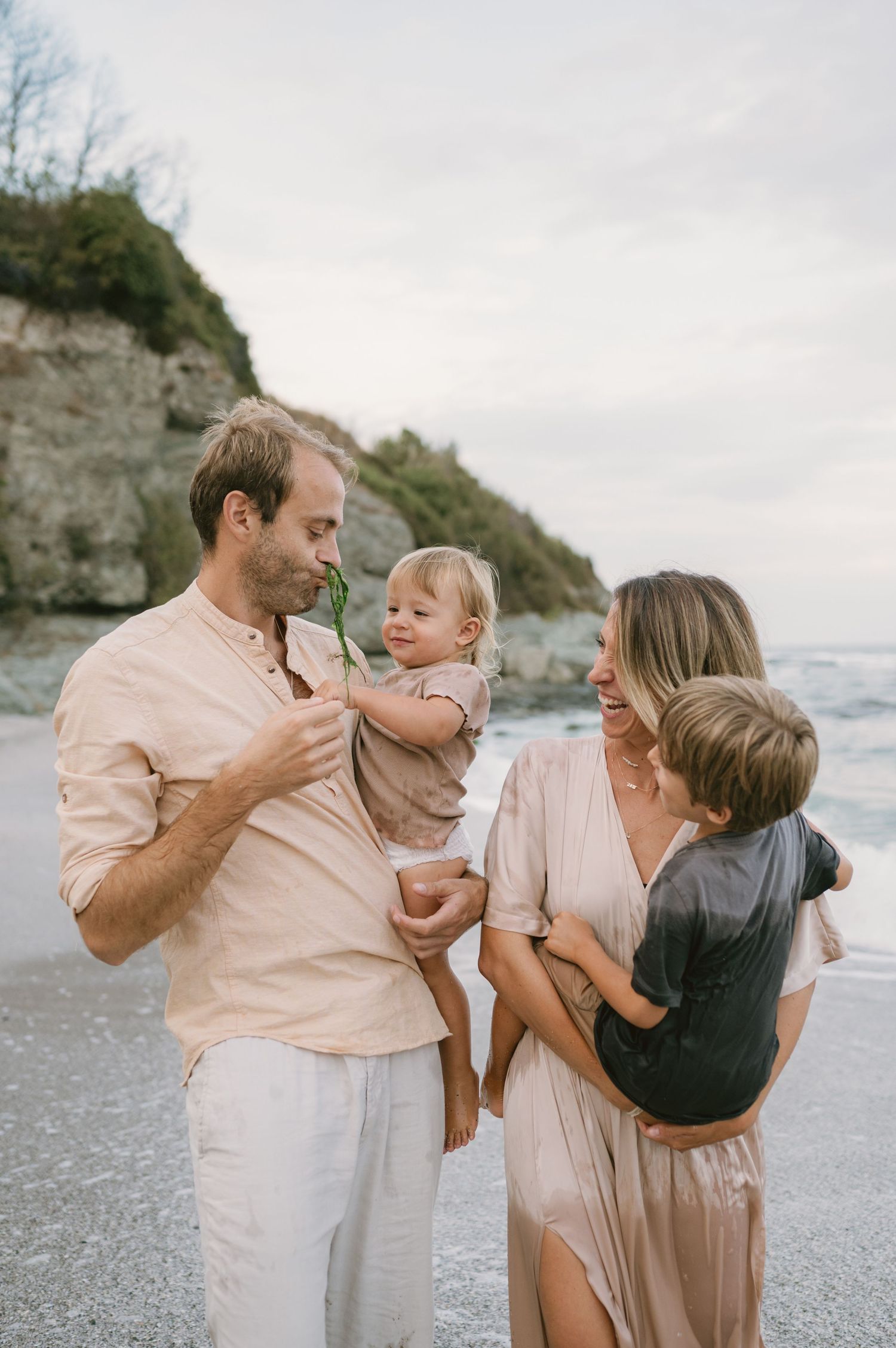 Family of four smiling and laughing together on a scenic beach, parents holding two young children near rocky cliffs.