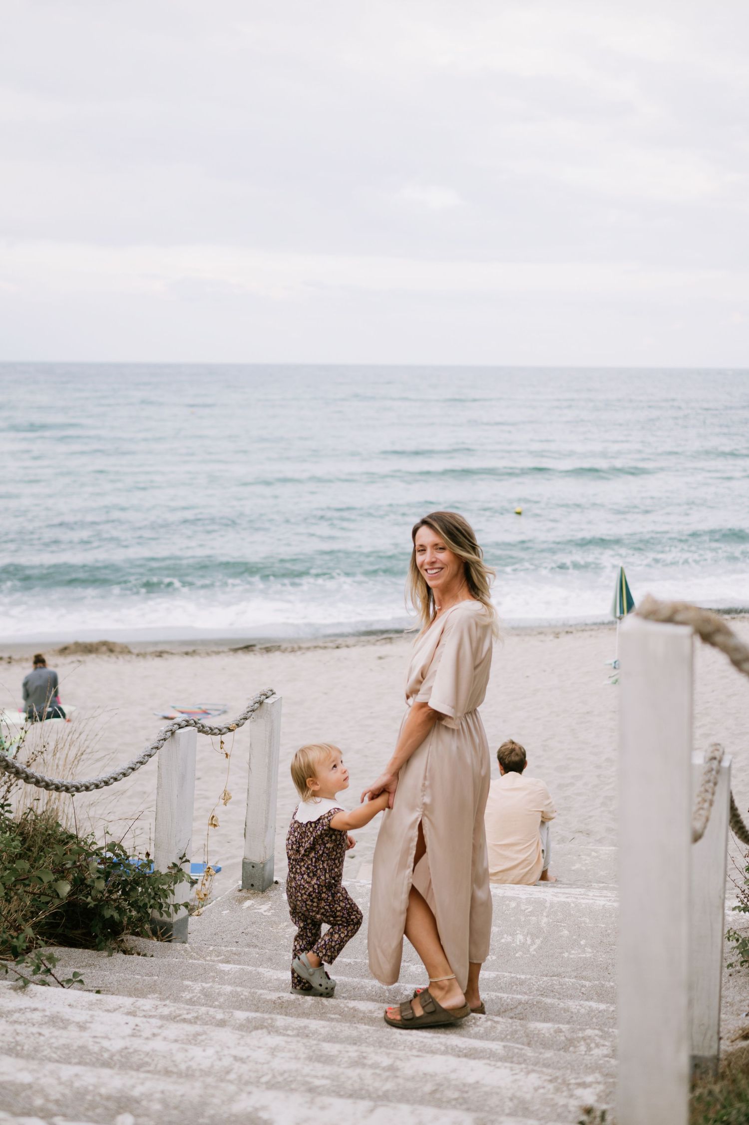 A woman in a beige dress holds a toddler's hand on beach stairs by the ocean on a cloudy day.