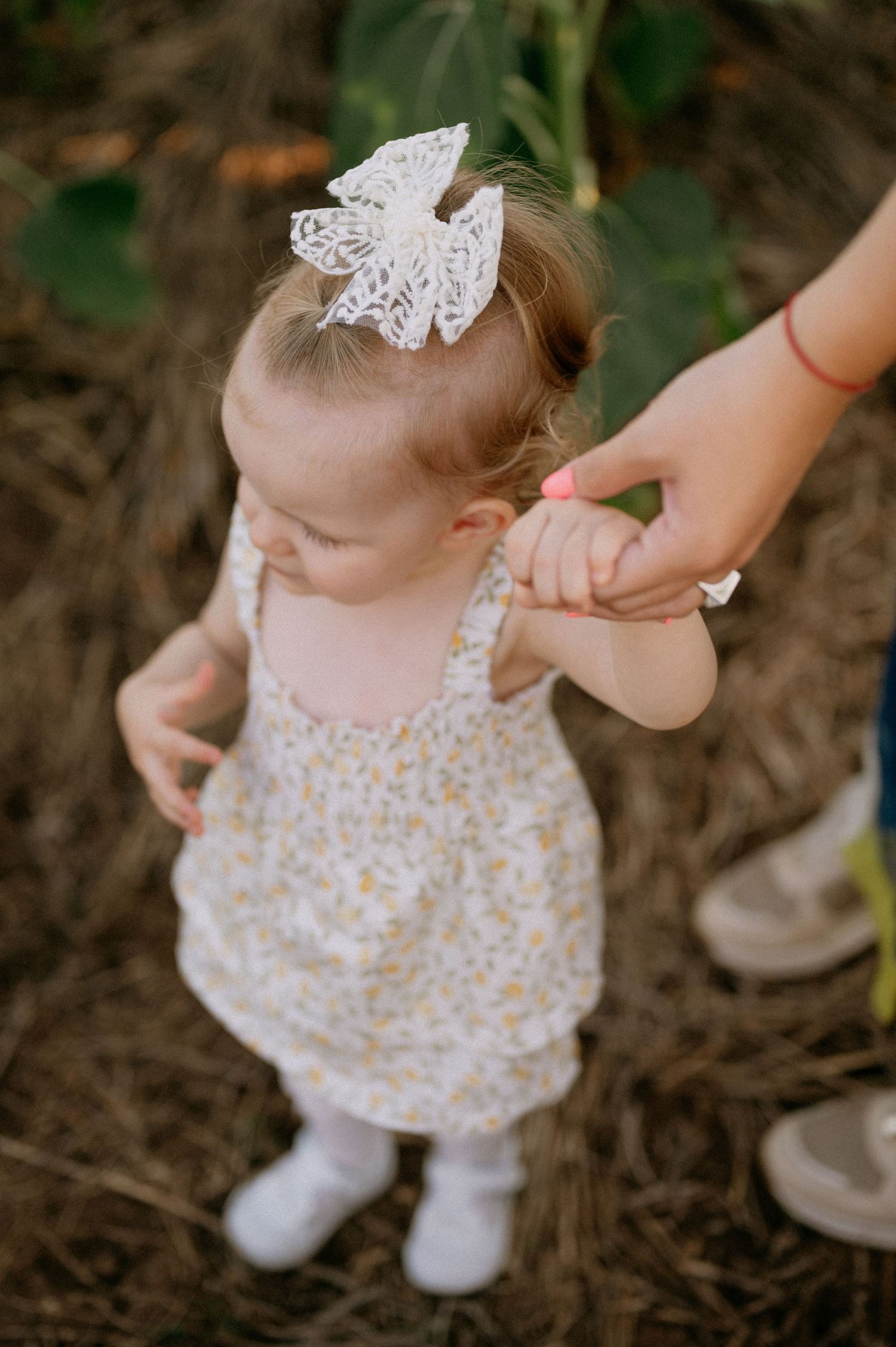 A toddler girl in a floral sundress with a white lace bow holds an adult's hand outdoors.