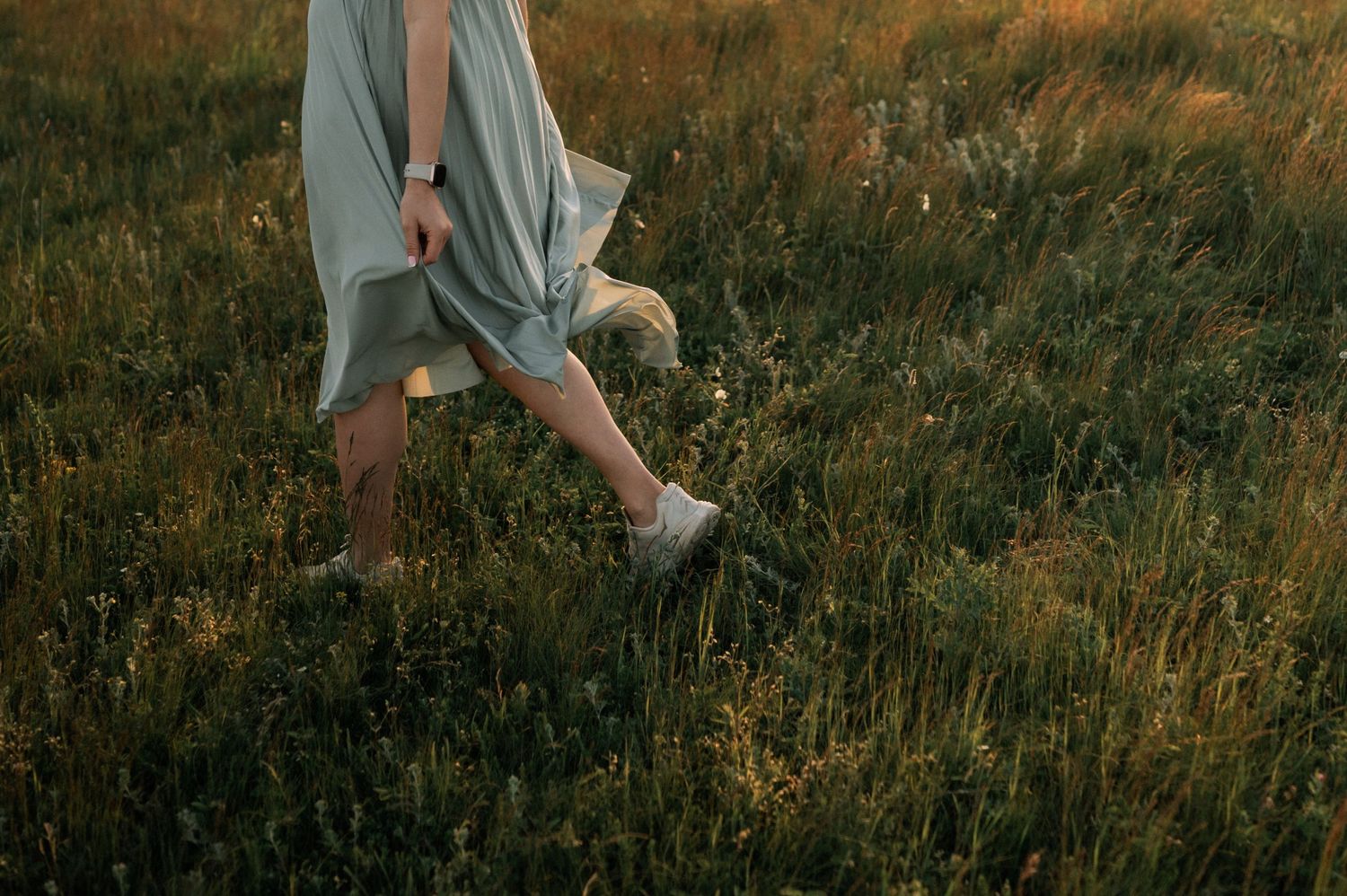 A woman in a light blue dress walks barefoot through a lush green meadow at golden hour sunset.
