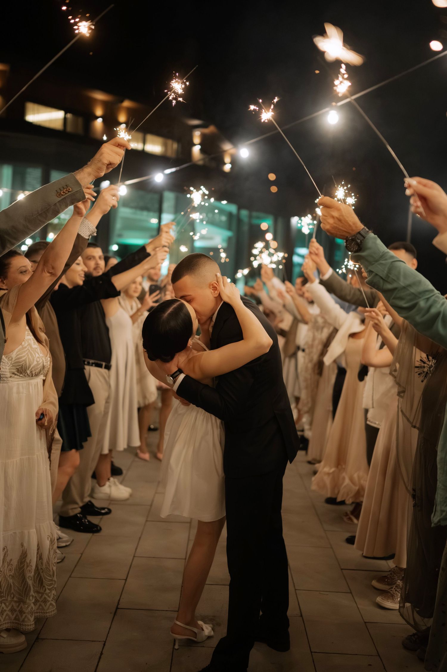 Wedding guests hold sparklers during an evening celebration on a patio with string lights overhead.
