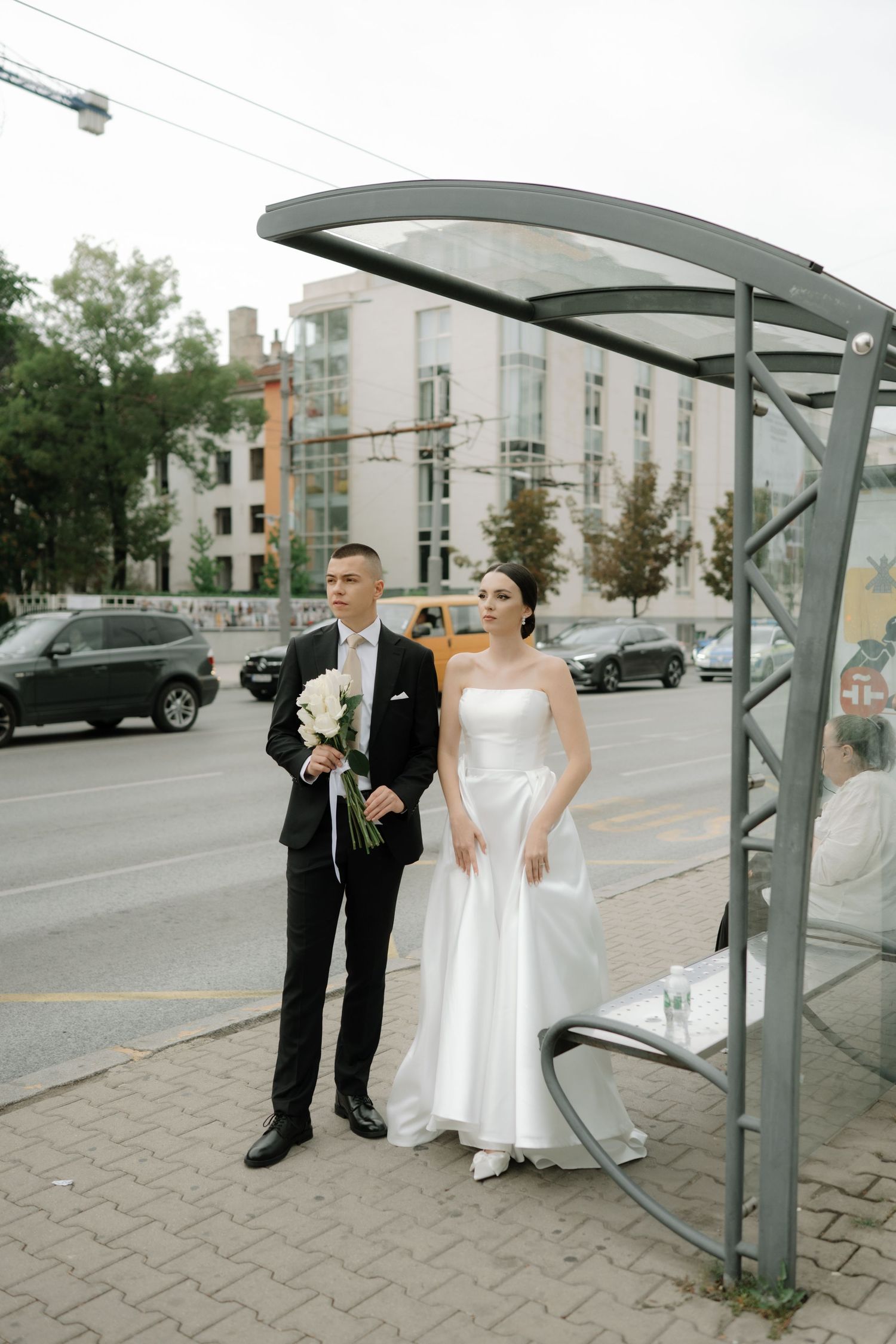 A couple in formal wedding attire poses playfully at a modern bus shelter on their special day.