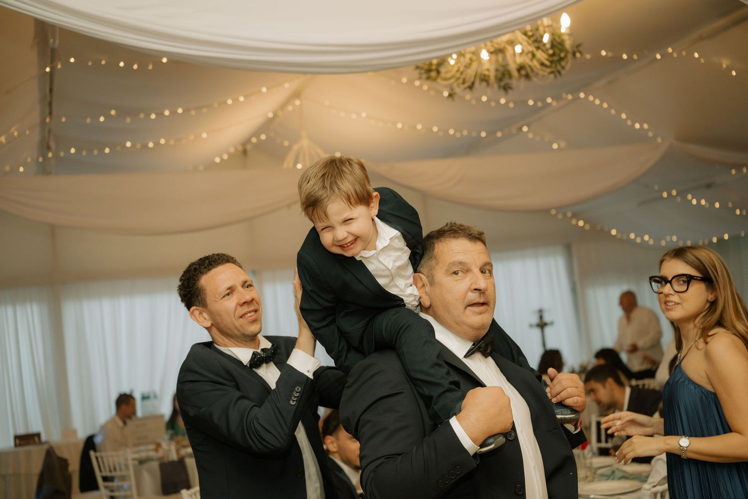 Young boy sits on man's shoulders laughing at wedding reception with fairy lights and draped ceiling.