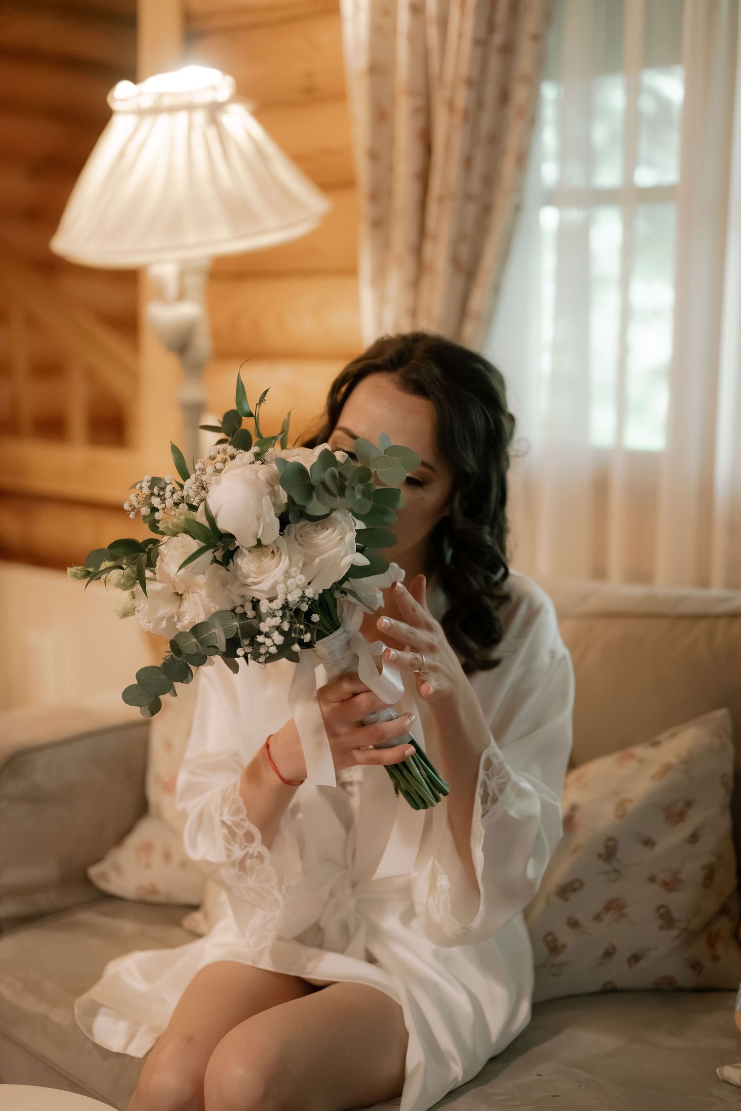 Bride in white lace robe holding elegant bouquet of white roses and eucalyptus while sitting on a couch.