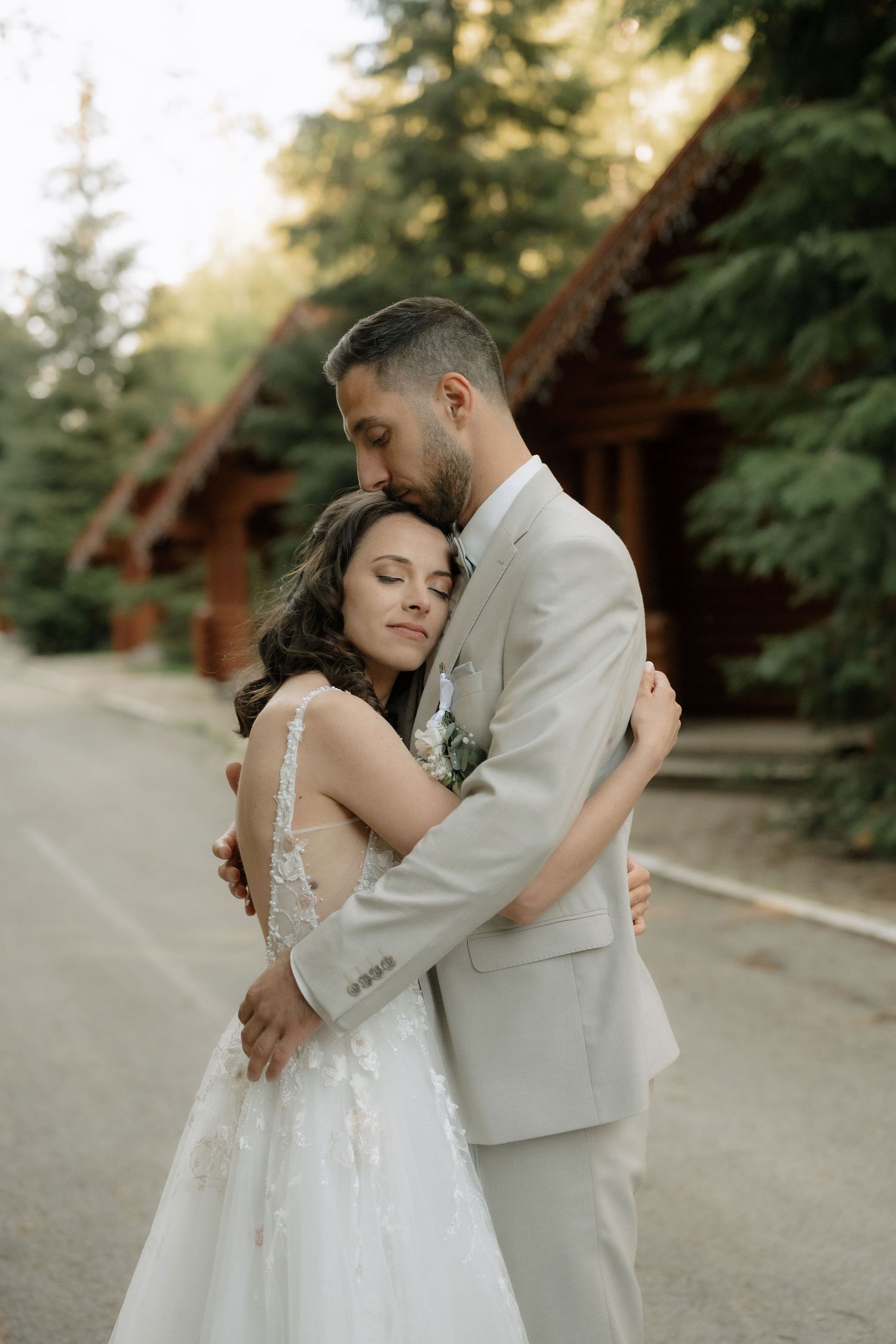 A groom in a gray suit tenderly embraces his bride in a white lace gown outdoors near rustic wooden cabins and pine trees.