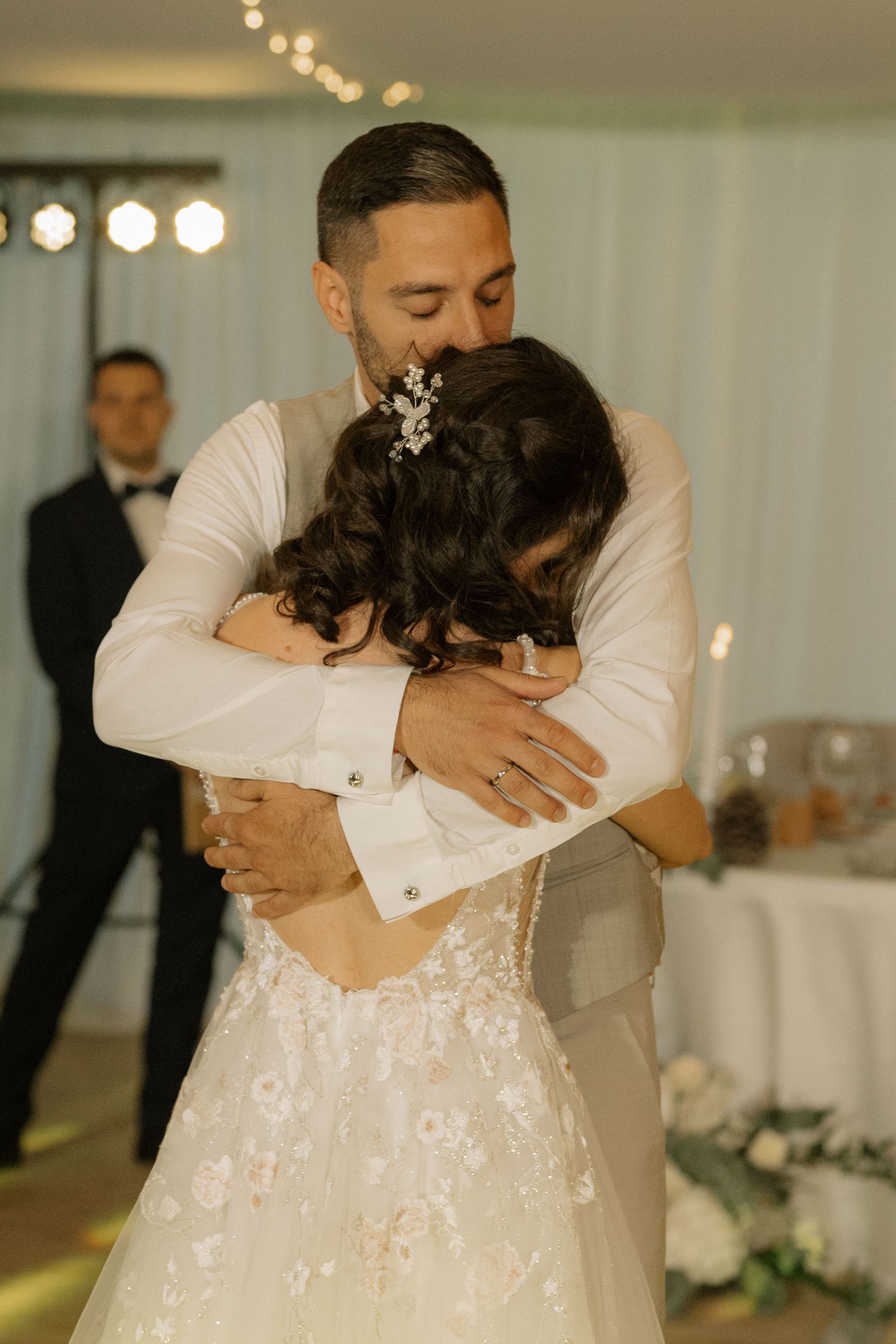 A groom in a white suit embraces his bride in a floral wedding gown during their first dance at a reception.