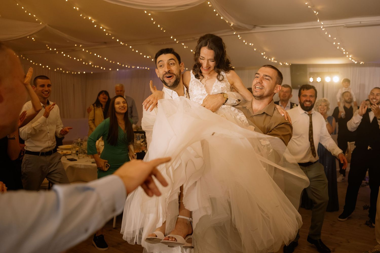 Joyful wedding reception dance with bride lifted by groomsmen, guests celebrating under fairy lights.