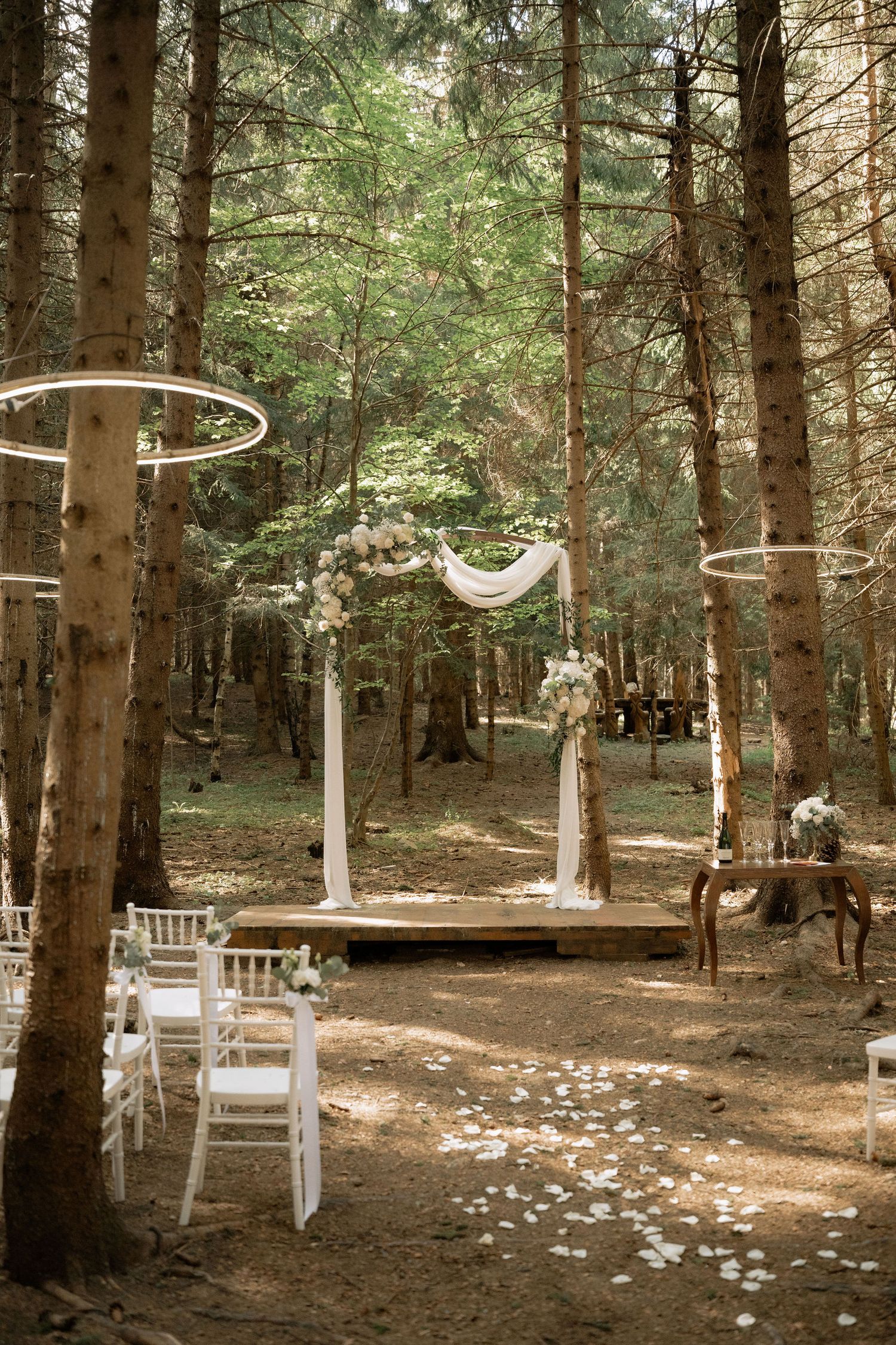 Enchanting forest wedding ceremony with white floral arch, draped fabric, Chiavari chairs, and hanging hoop decor.