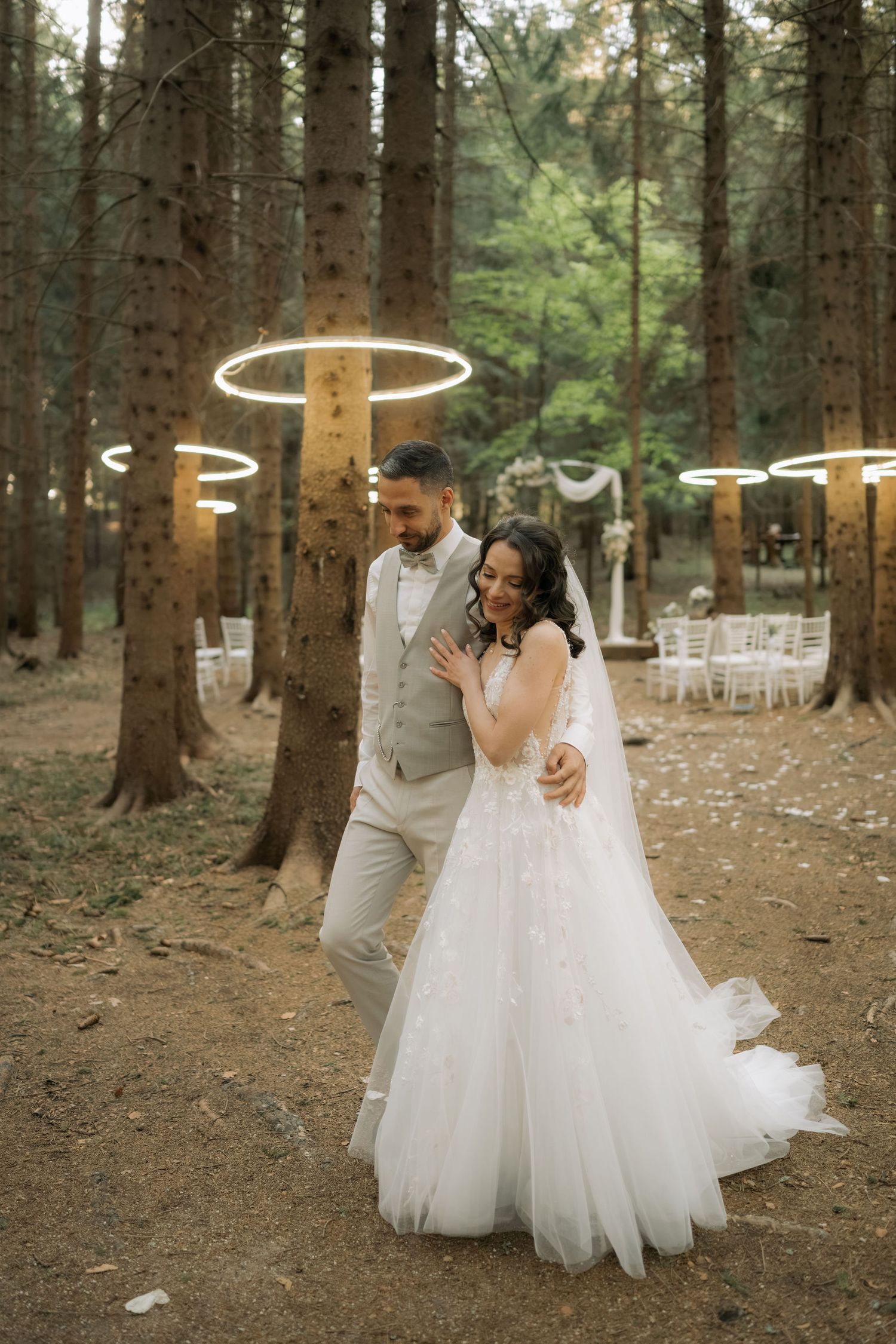 Bride and groom walking together in a forest wedding venue with glowing circular lights hanging from tall pine trees.