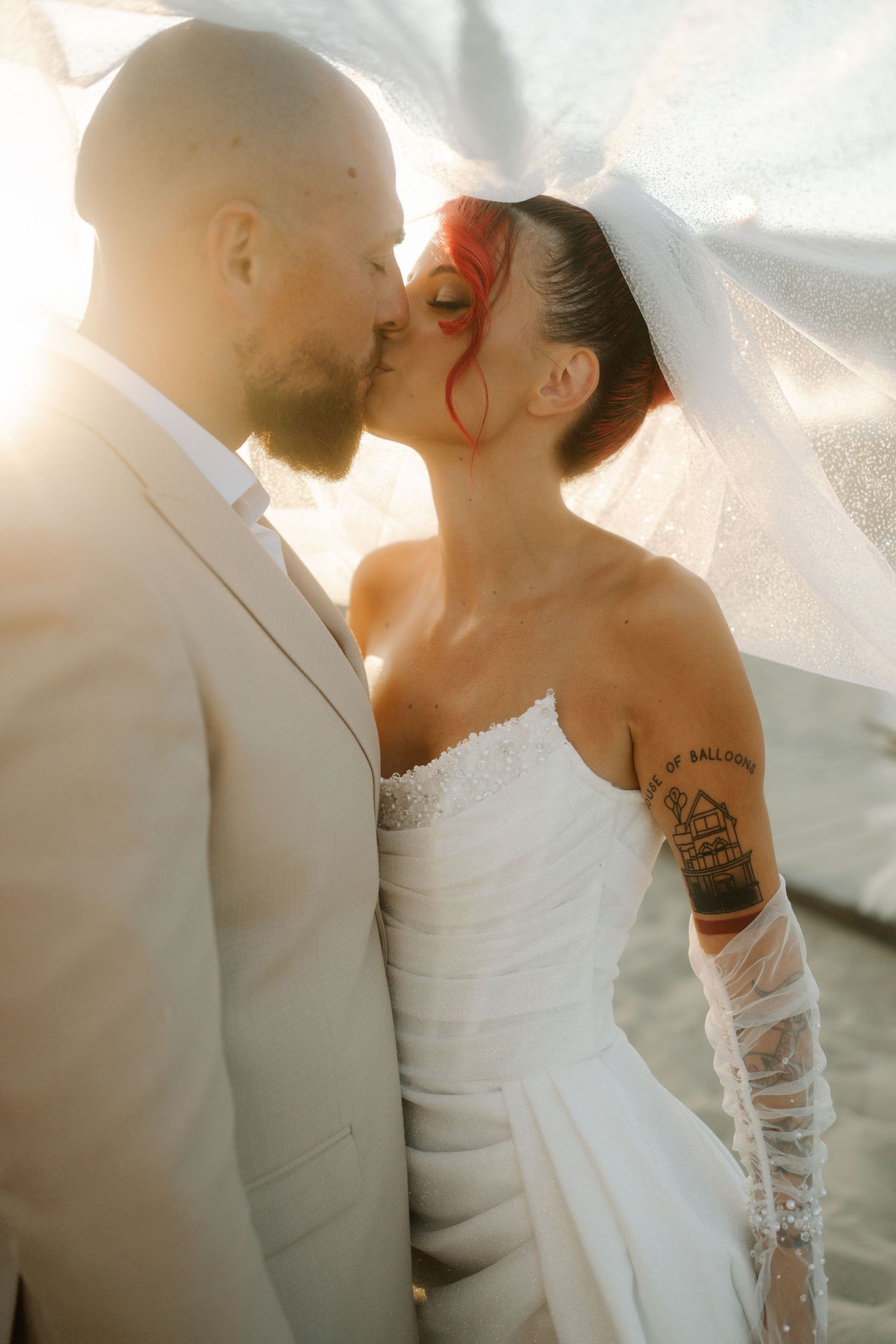 A romantic wedding couple shares a tender kiss in soft sunlight while wearing white formal attire.