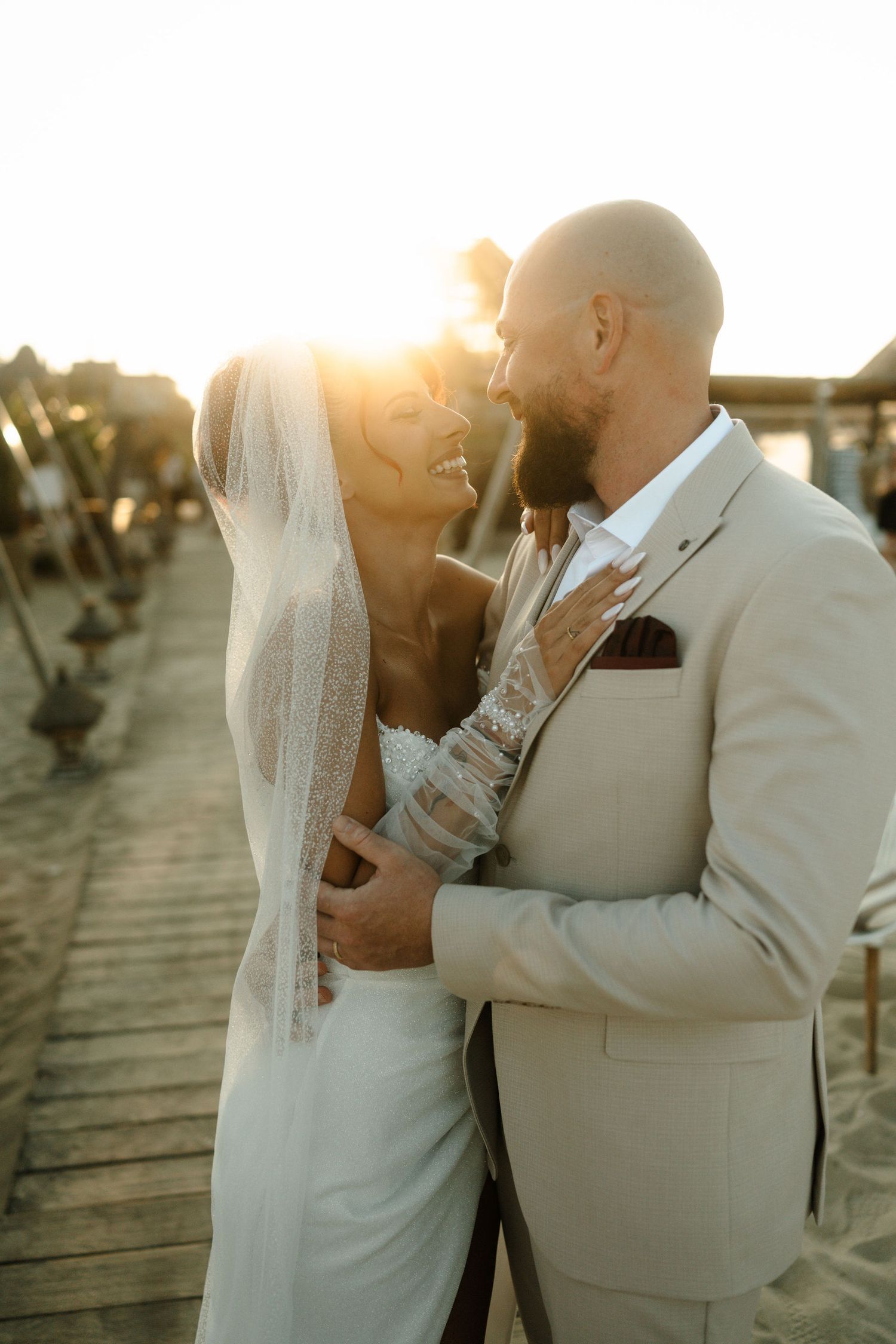 A couple in wedding attire embraces on a wooden boardwalk at sunset, creating a romantic silhouette against golden light.