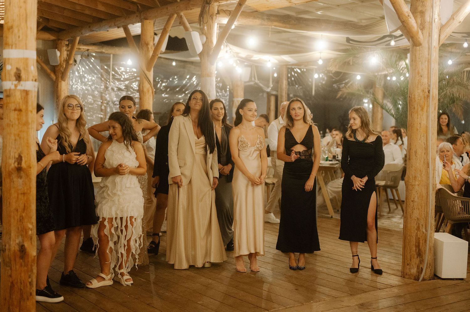 Wedding guests line up against a rustic wooden wall decorated with string lights during a reception celebration.