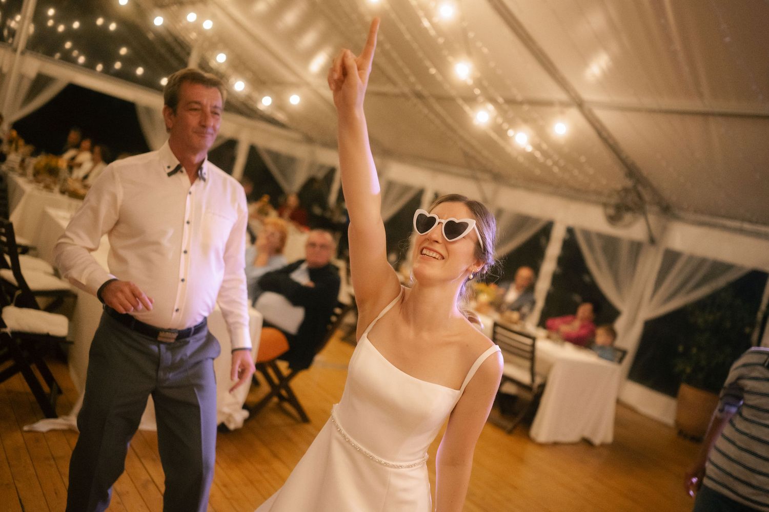Wedding guests dance and celebrate on a wooden dance floor under twinkling lights at an indoor reception venue.
