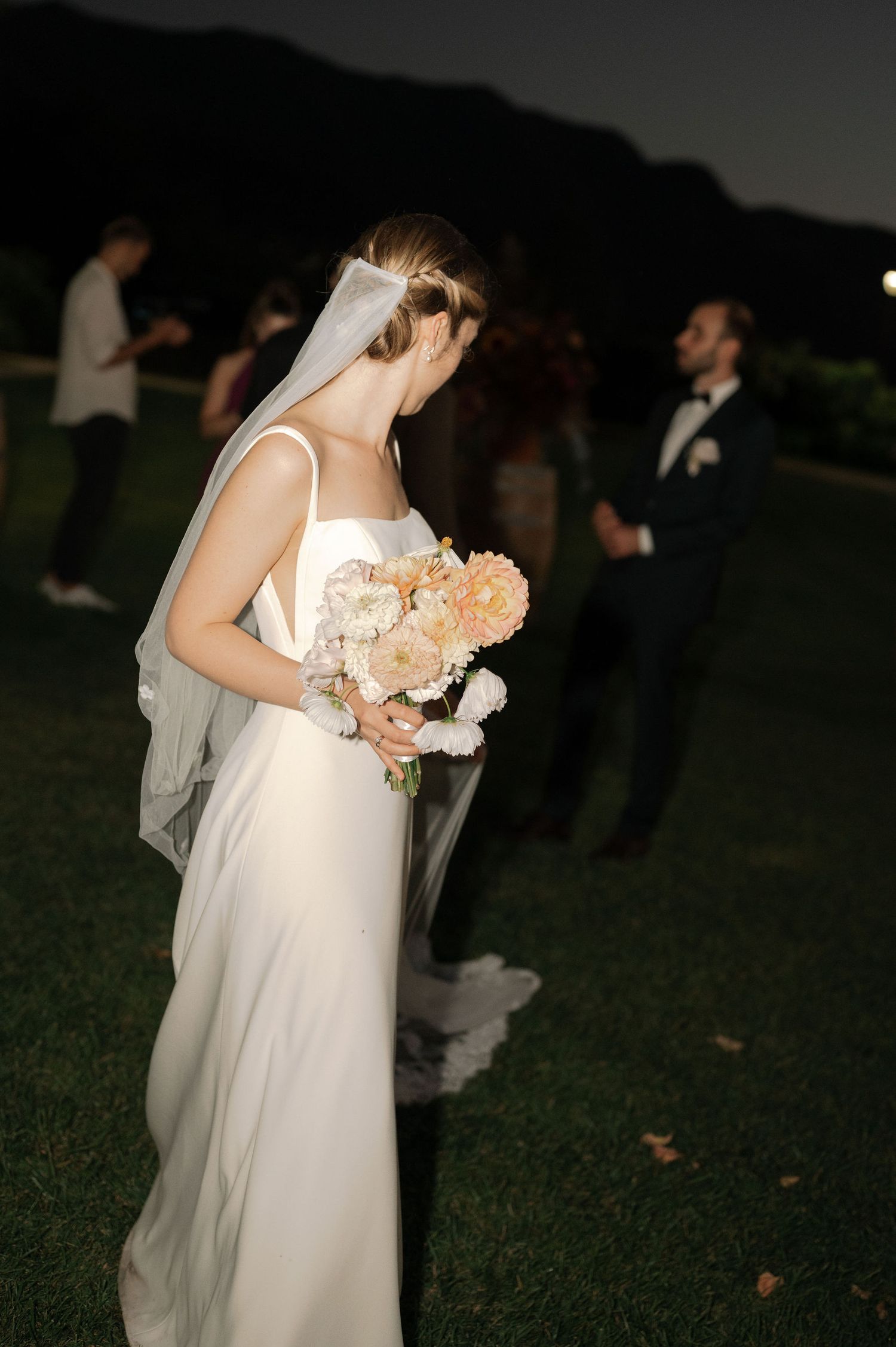 A bride in a flowing white dress holds a bouquet of peach and white flowers at an outdoor evening wedding ceremony.