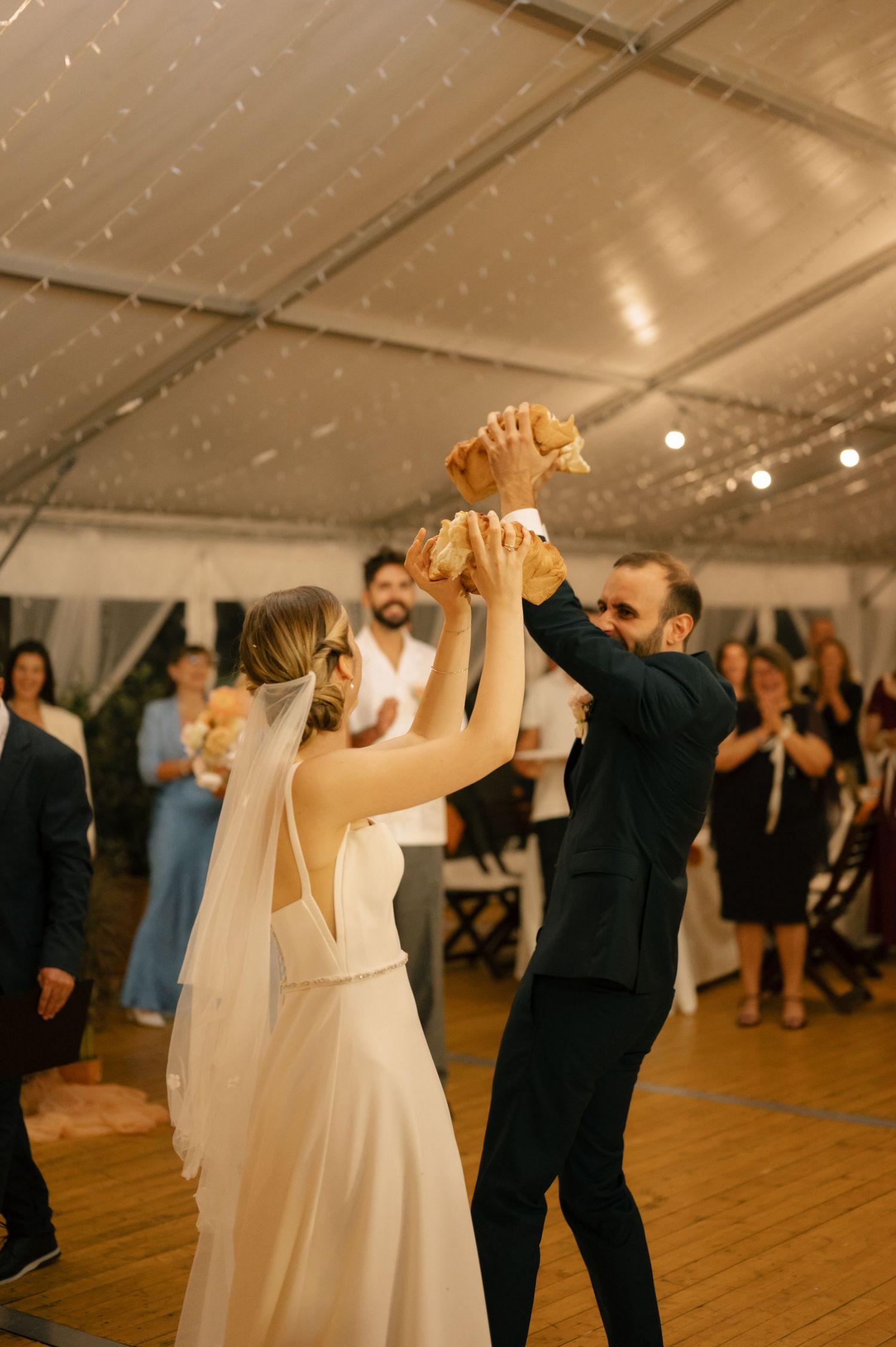 Wedding guests dancing in pairs on a wooden dance floor under a white tent with string lights.