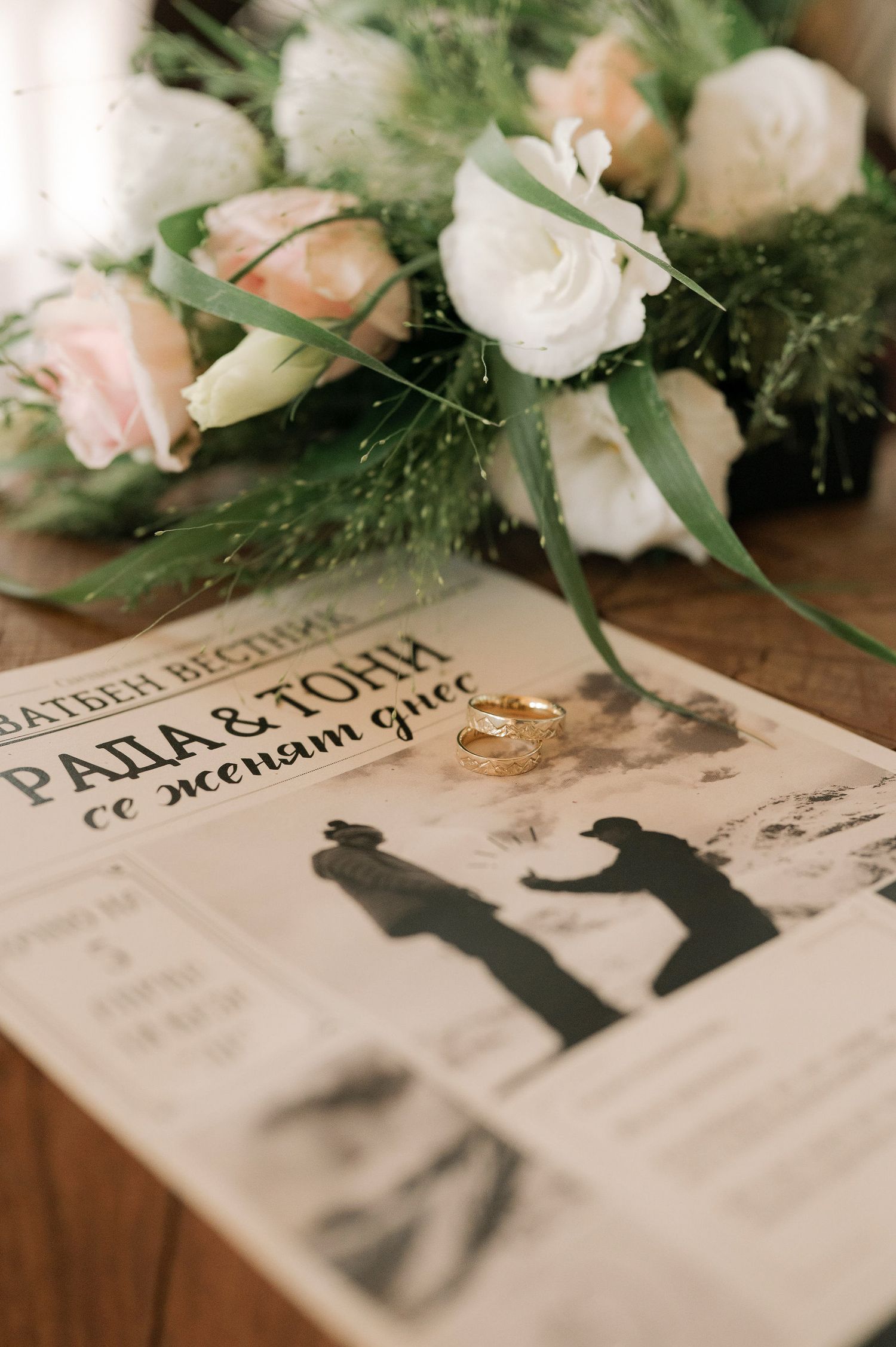 Wedding invitation with silhouettes lies on wooden surface surrounded by delicate pink and white flowers and greenery.