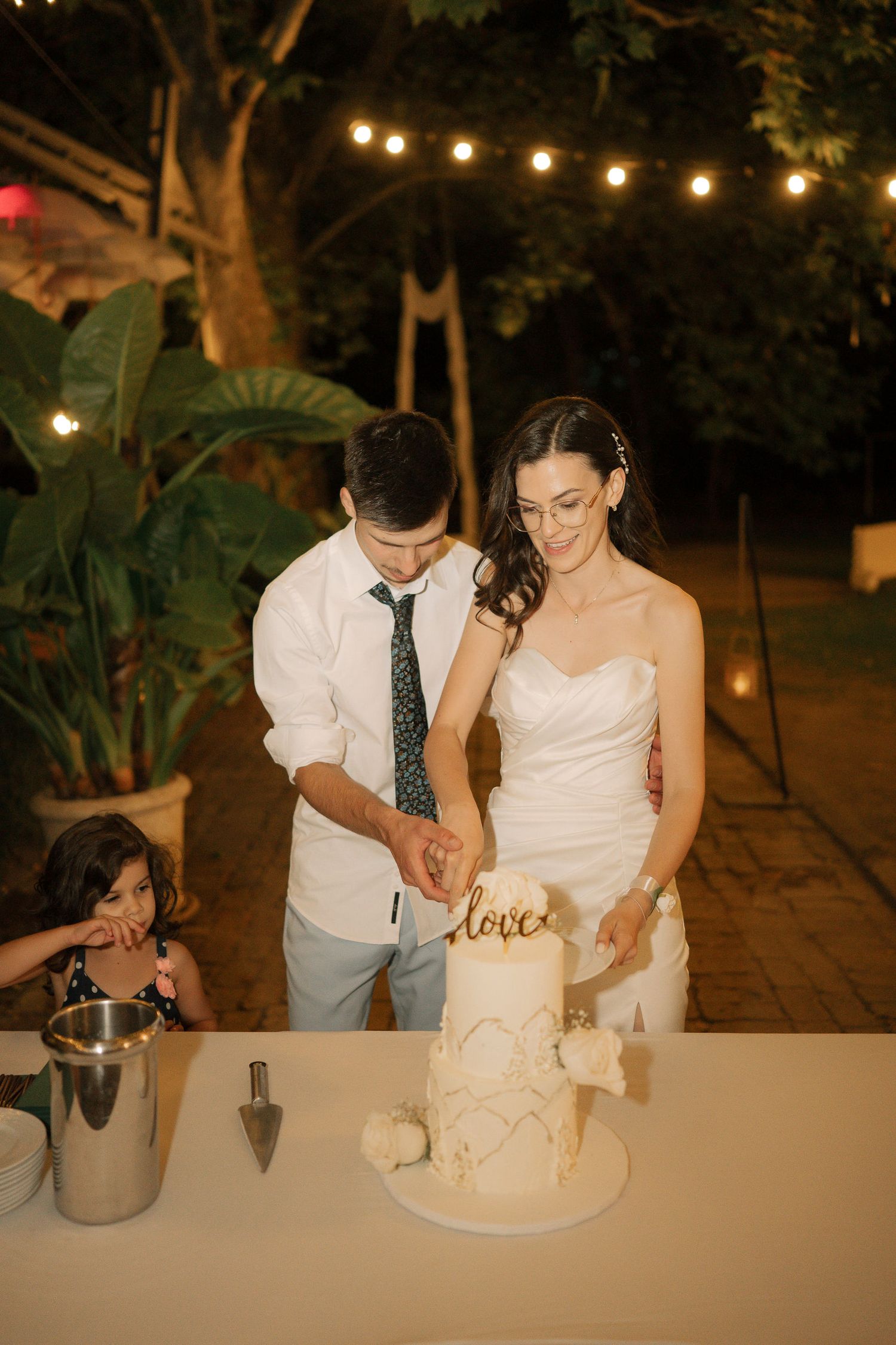 A couple cuts their elegant white wedding cake together under string lights at an outdoor evening reception.