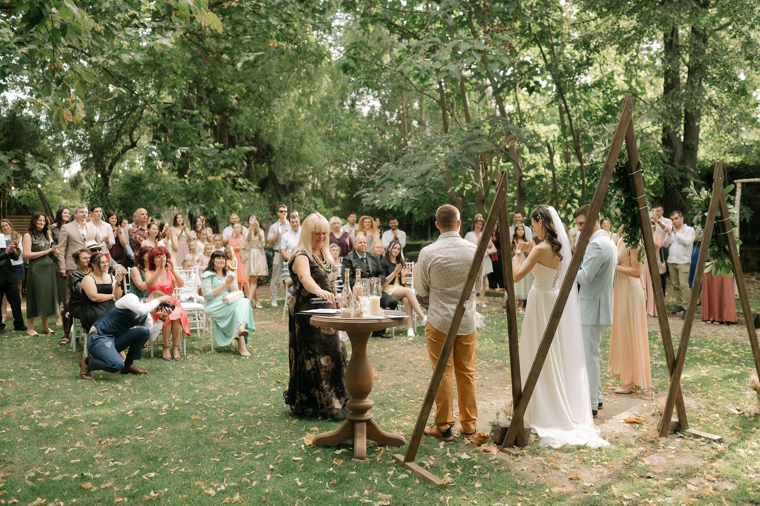 Wedding guests gather in a wooded outdoor setting for a celebration among trees and natural greenery.