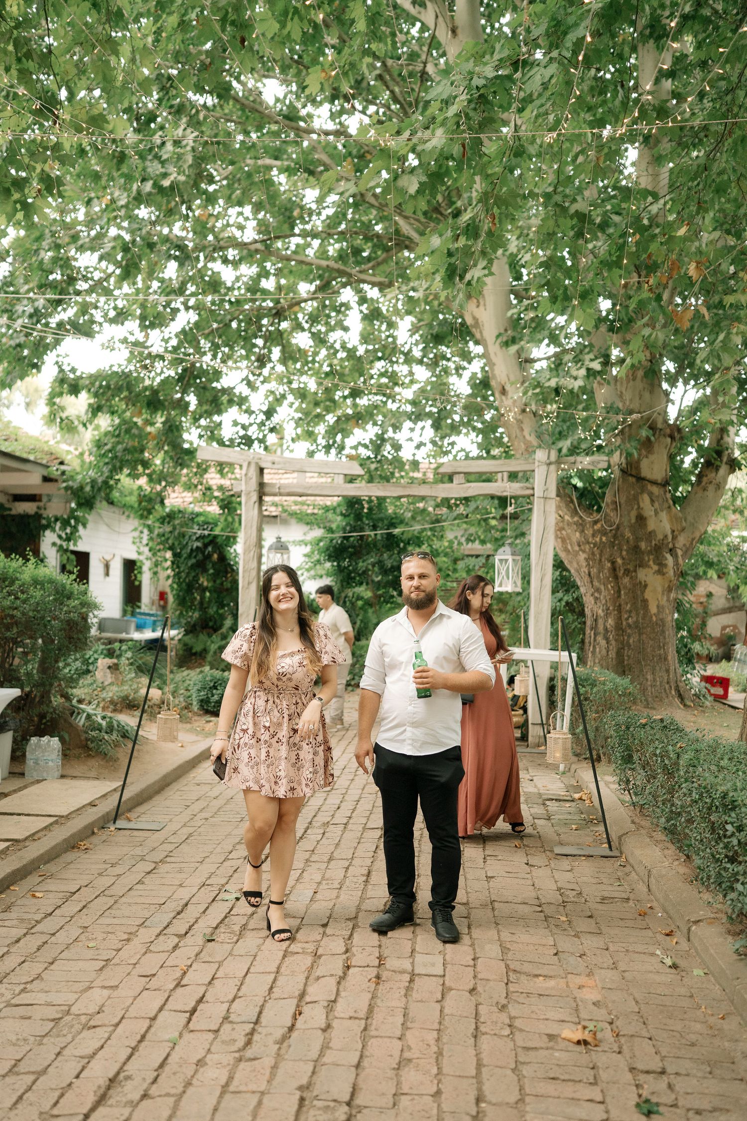 Two people walking together on a brick pathway surrounded by lush green trees on a sunny day.