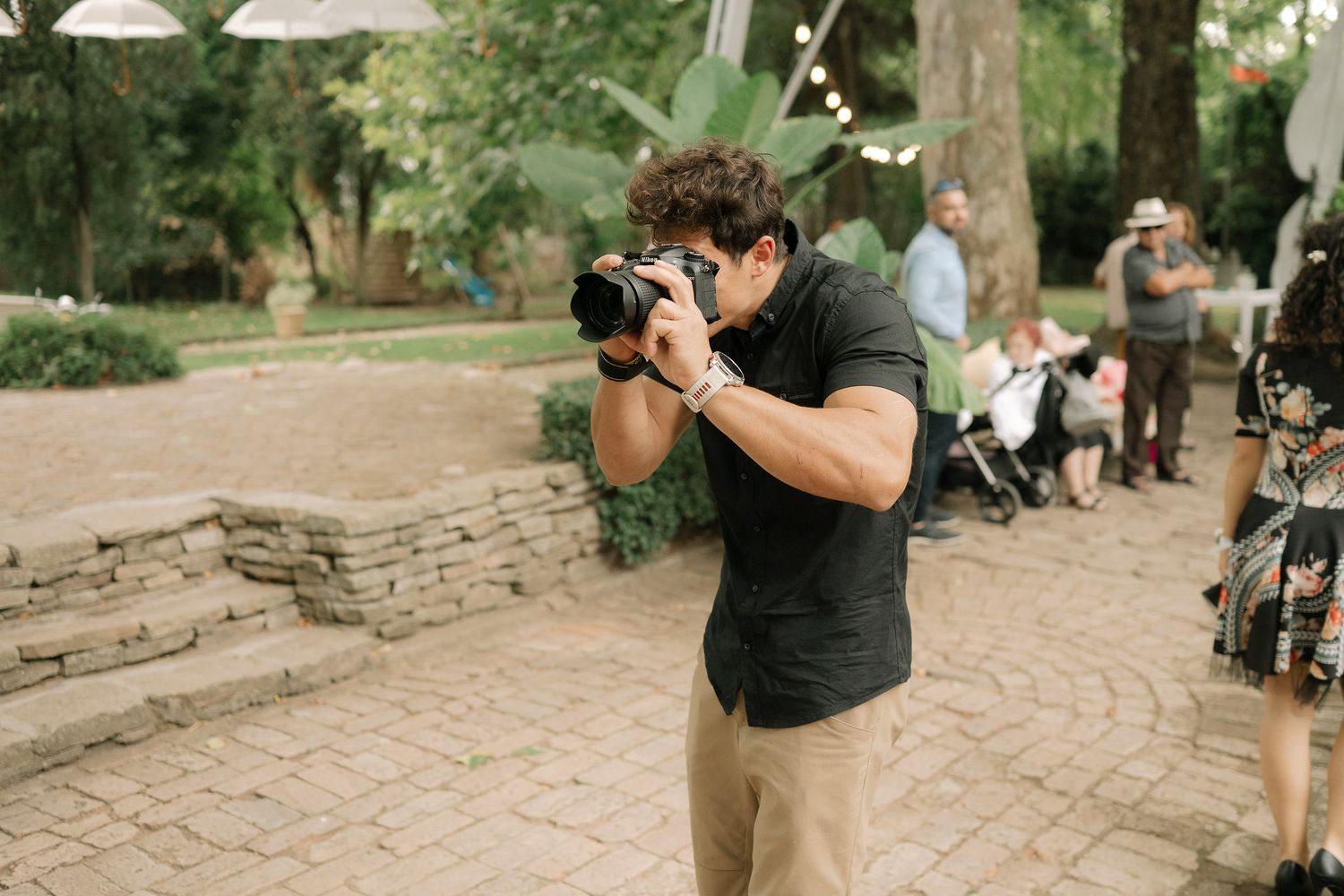 A photographer wearing a black shirt and khaki pants takes pictures on a brick pathway in a garden setting.