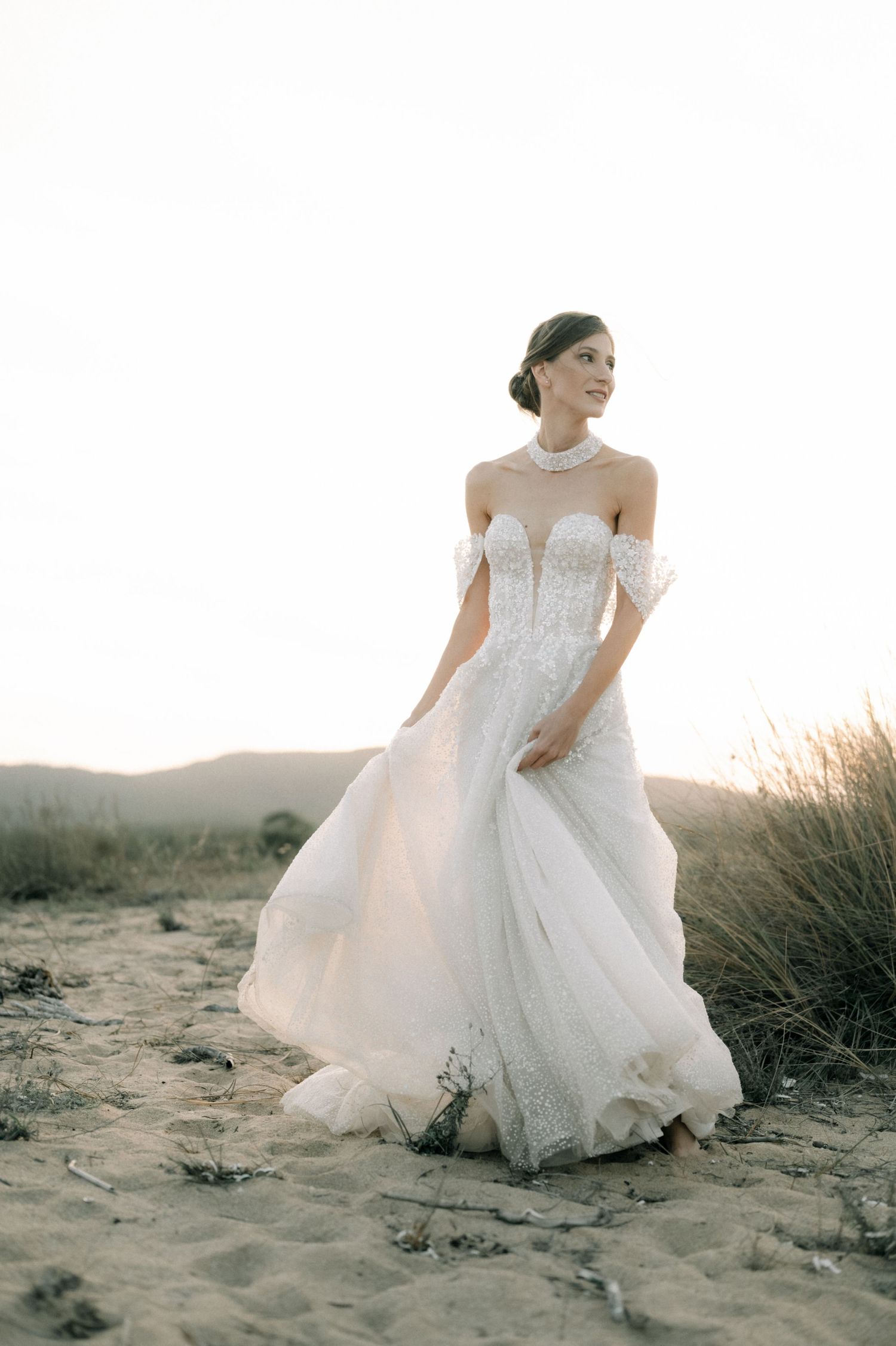 Bride in off-shoulder lace wedding gown standing on sandy beach at sunset, dress flowing in the breeze.