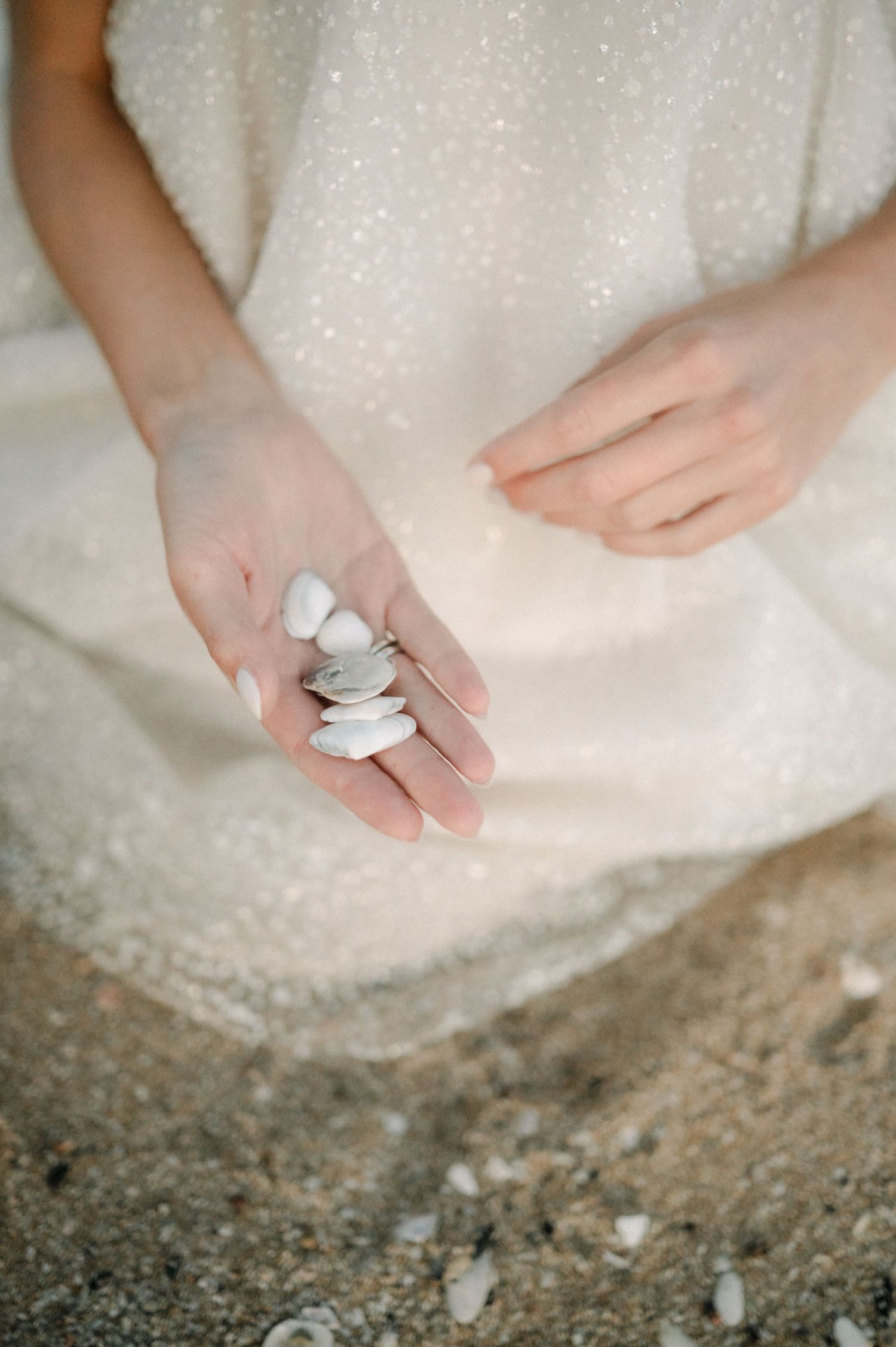 A woman in a white sparkly dress holds small white pebbles and shells in her open palm on a sandy beach.