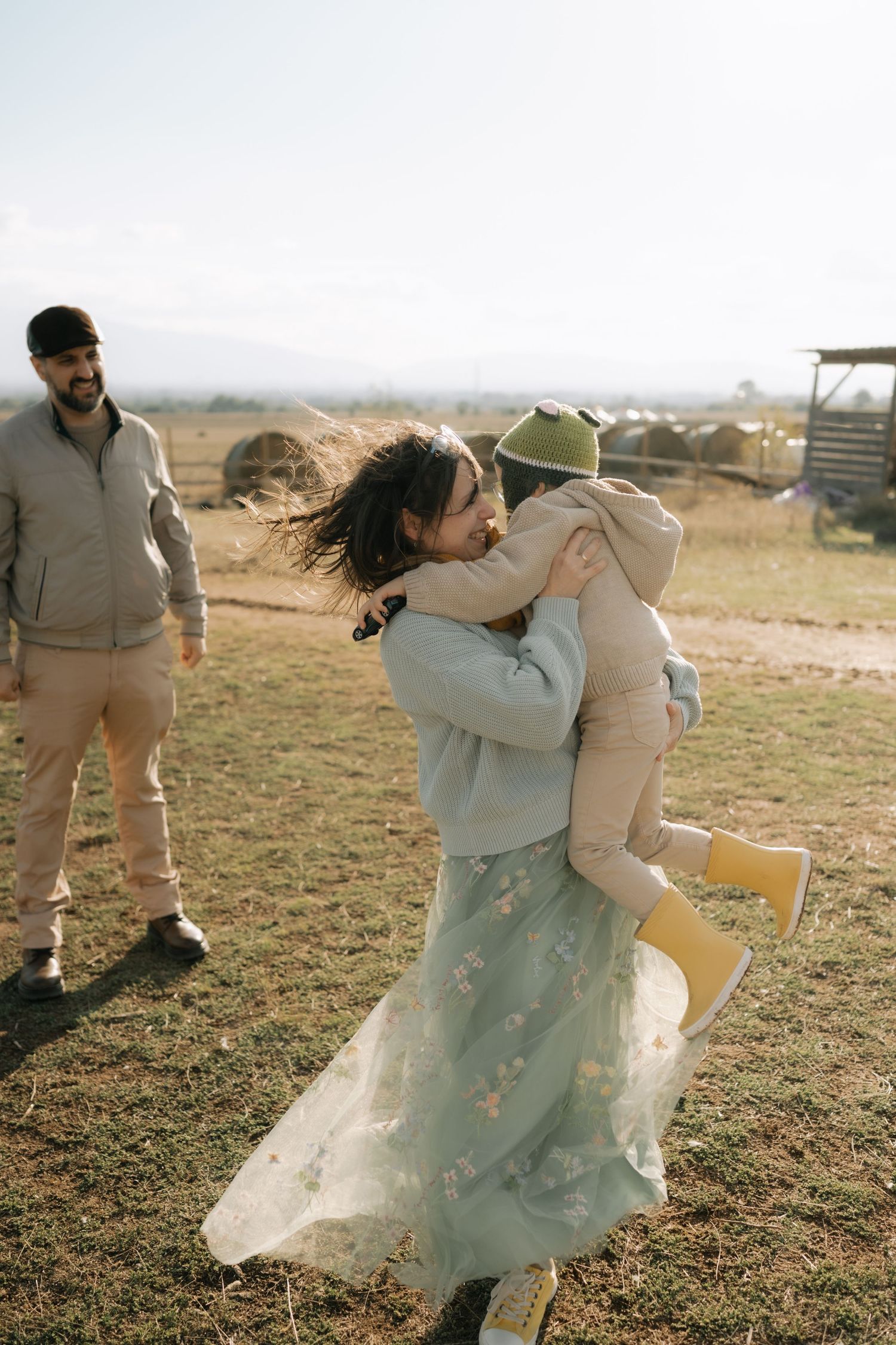 A tender family moment captured outdoors on a sunny day in a rustic countryside setting with flowing dresses and casual attire.