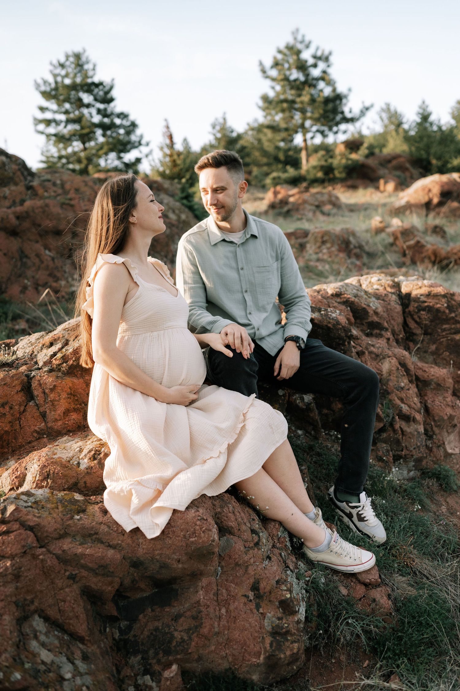 Couple sitting on rocky outcrop during sunset maternity photoshoot in nature with evergreen trees in background.