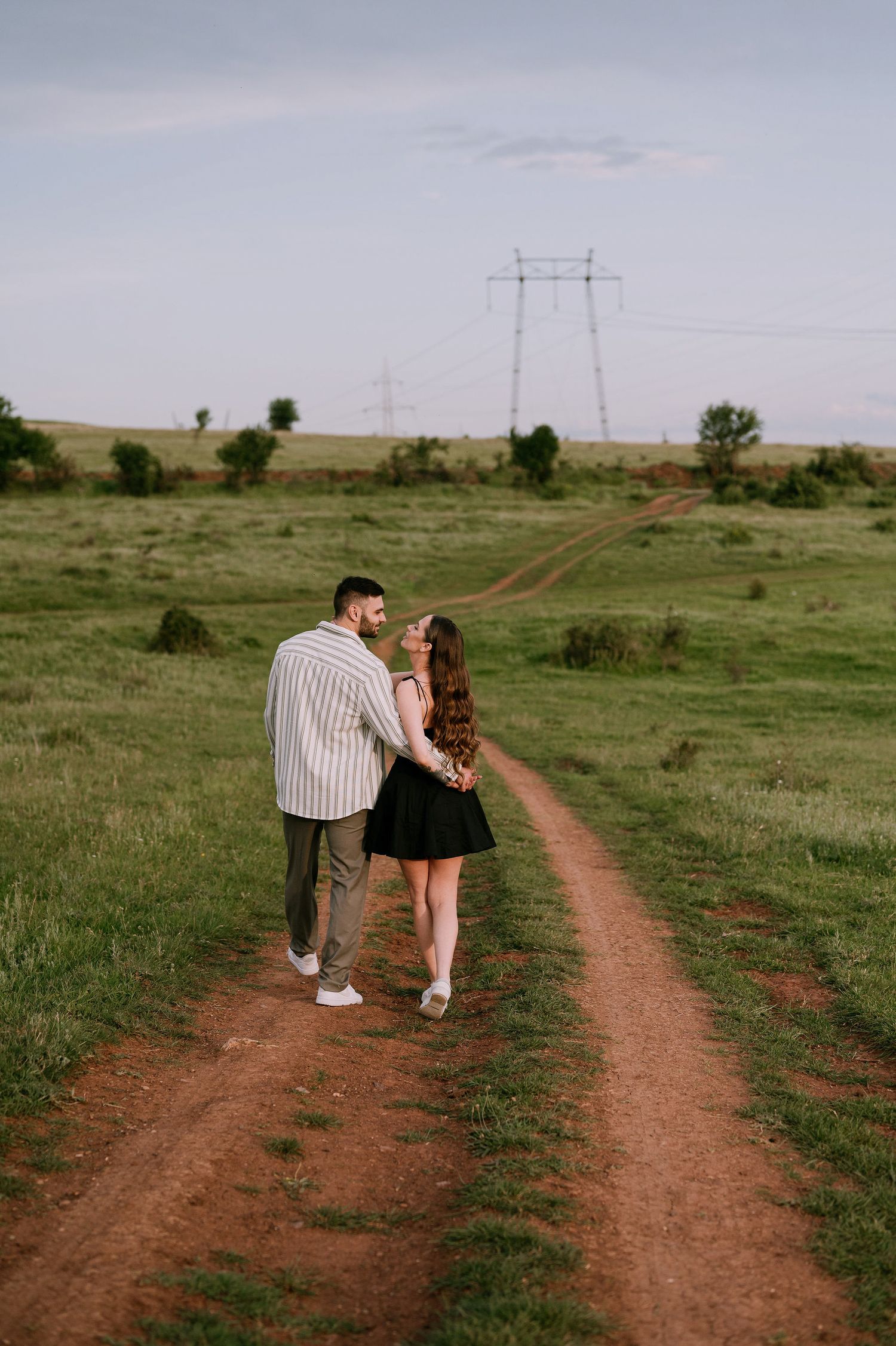 A couple walks together on a dirt path through a grassy field with power lines visible in the distance.