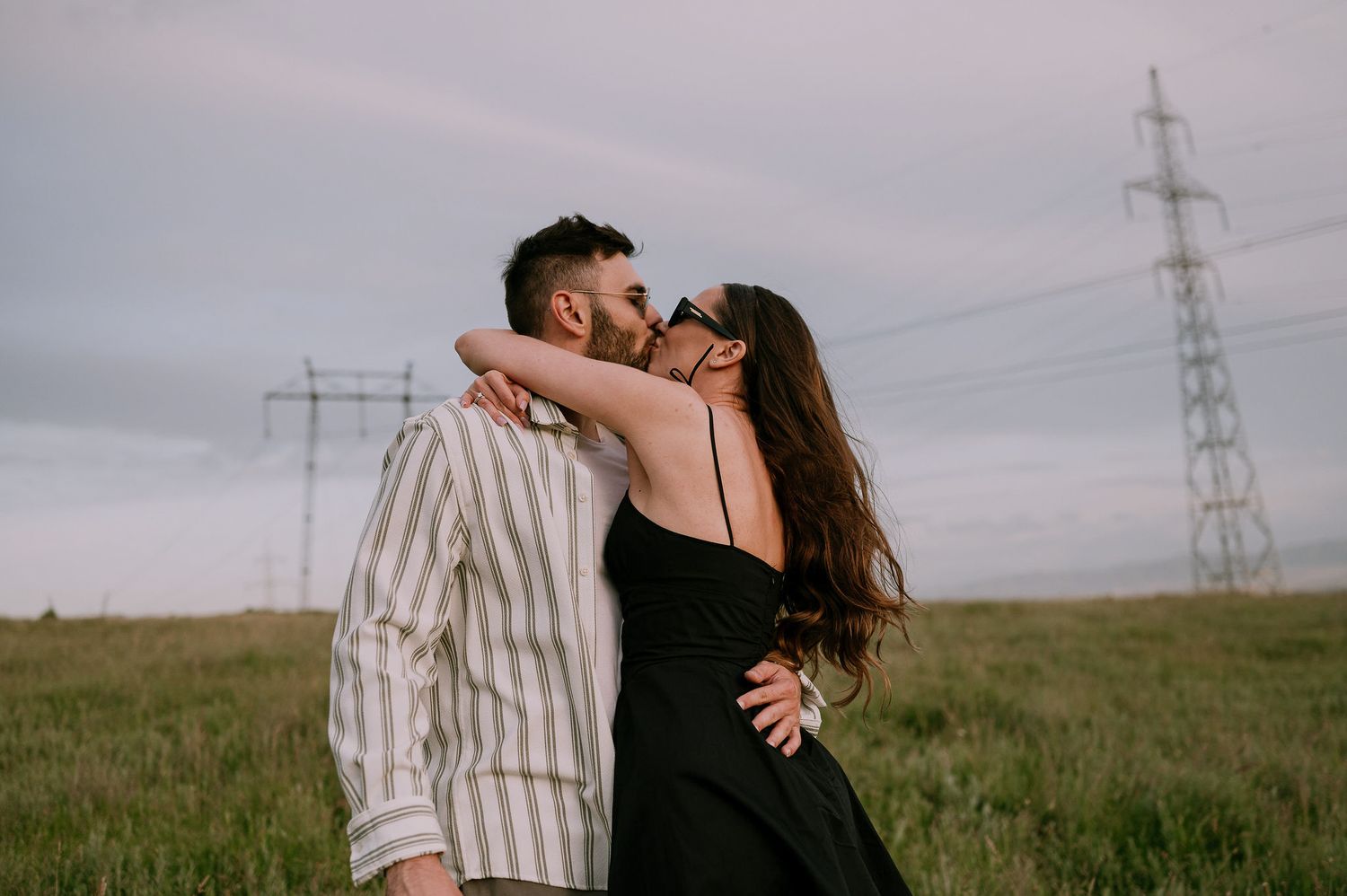 Romantic couple embraces in a grassy field with power lines and moody skies in the background.