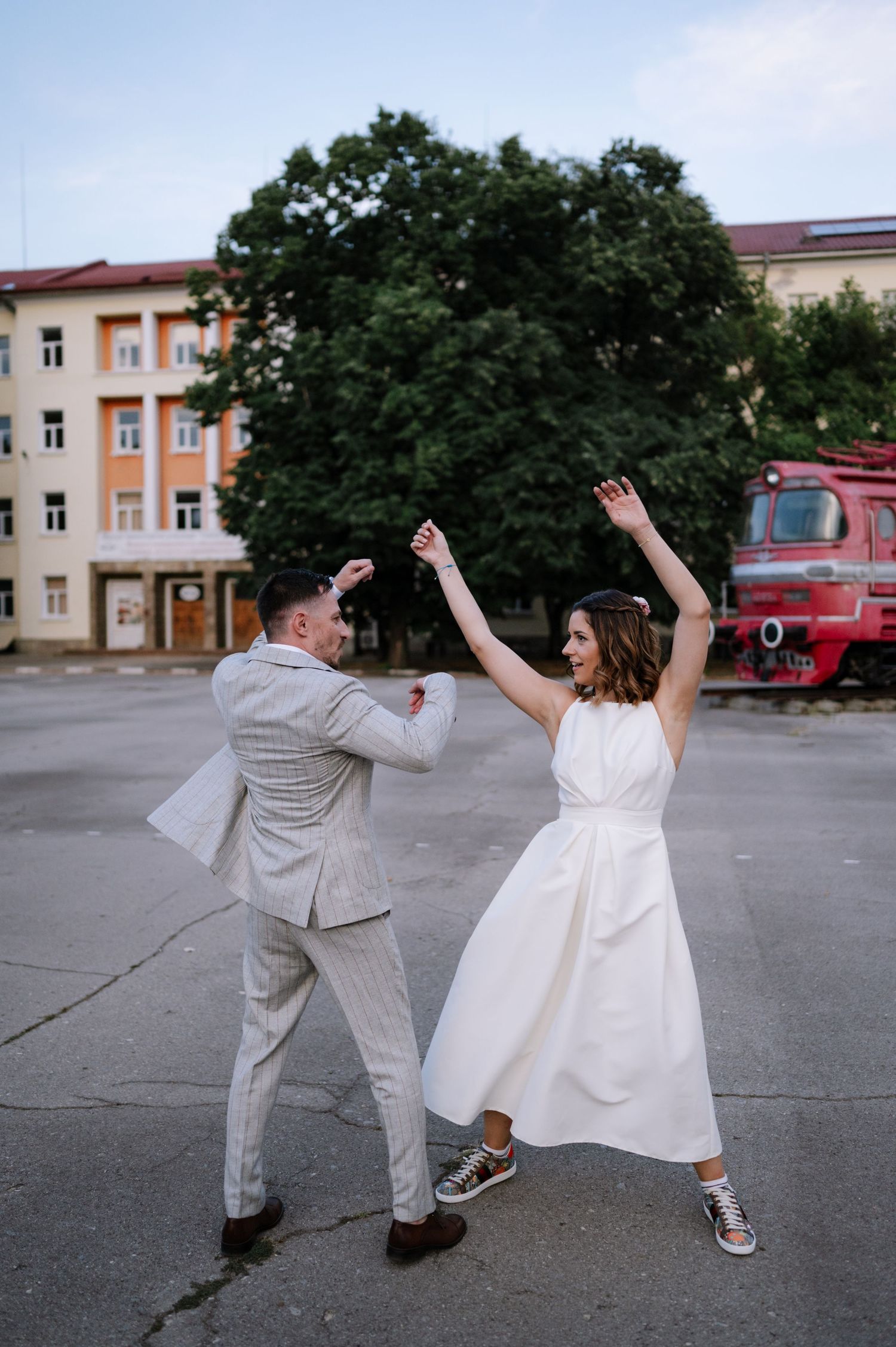 A couple in wedding attire playfully dances and poses in a parking lot with buildings in the background.