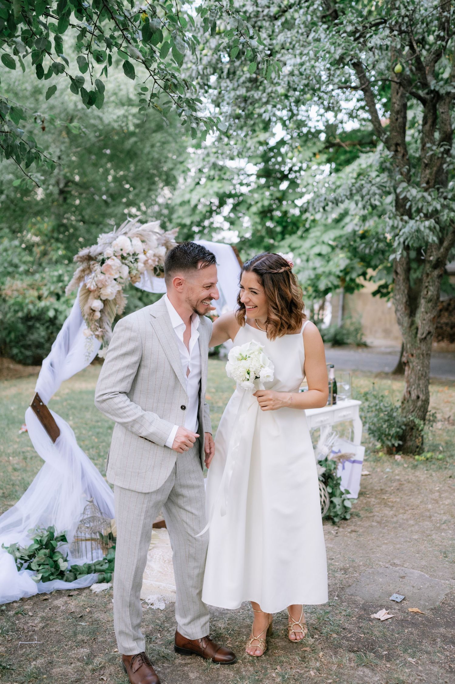 A wedding party in light colored attire poses playfully together in a wooded outdoor setting.