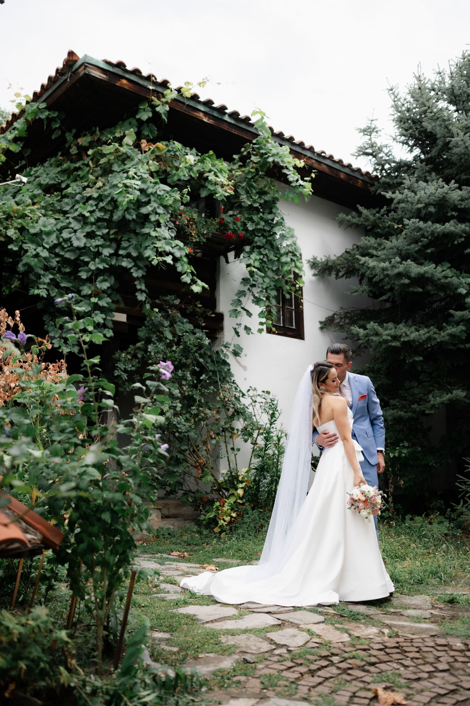 A romantic wedding couple shares an intimate moment in a garden path surrounded by lush greenery and white walls.