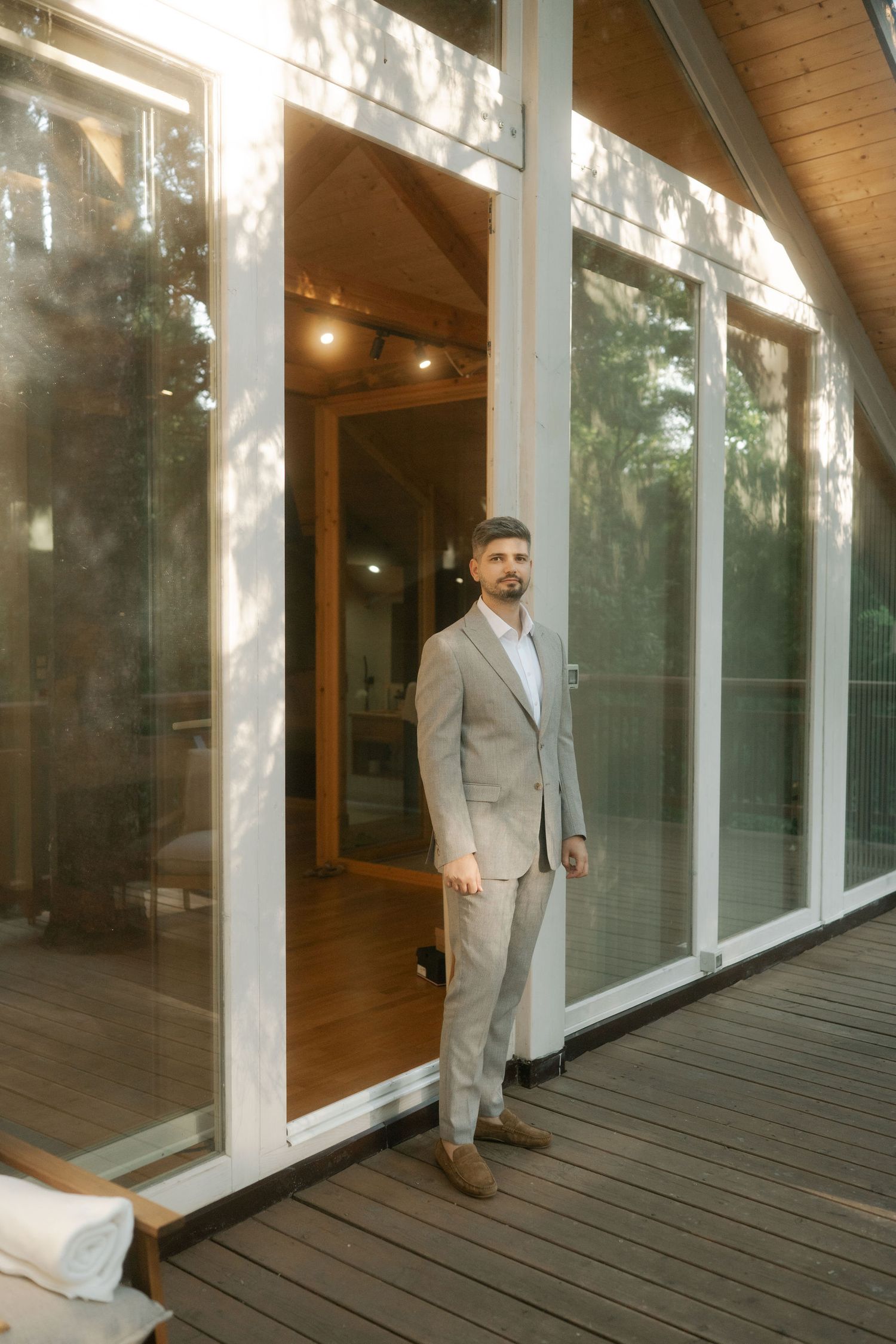 A person in a light gray suit stands on a wooden deck next to large glass windows of a modern building.