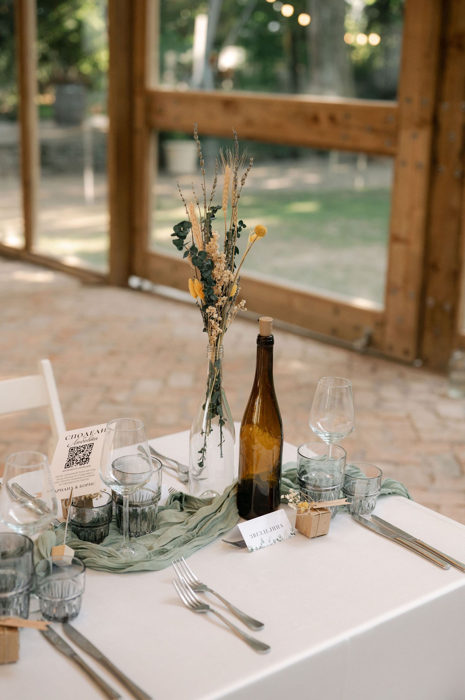 Close-up view of minimalist table decor featuring sage green fabric, wine bottles, and place settings in natural light.