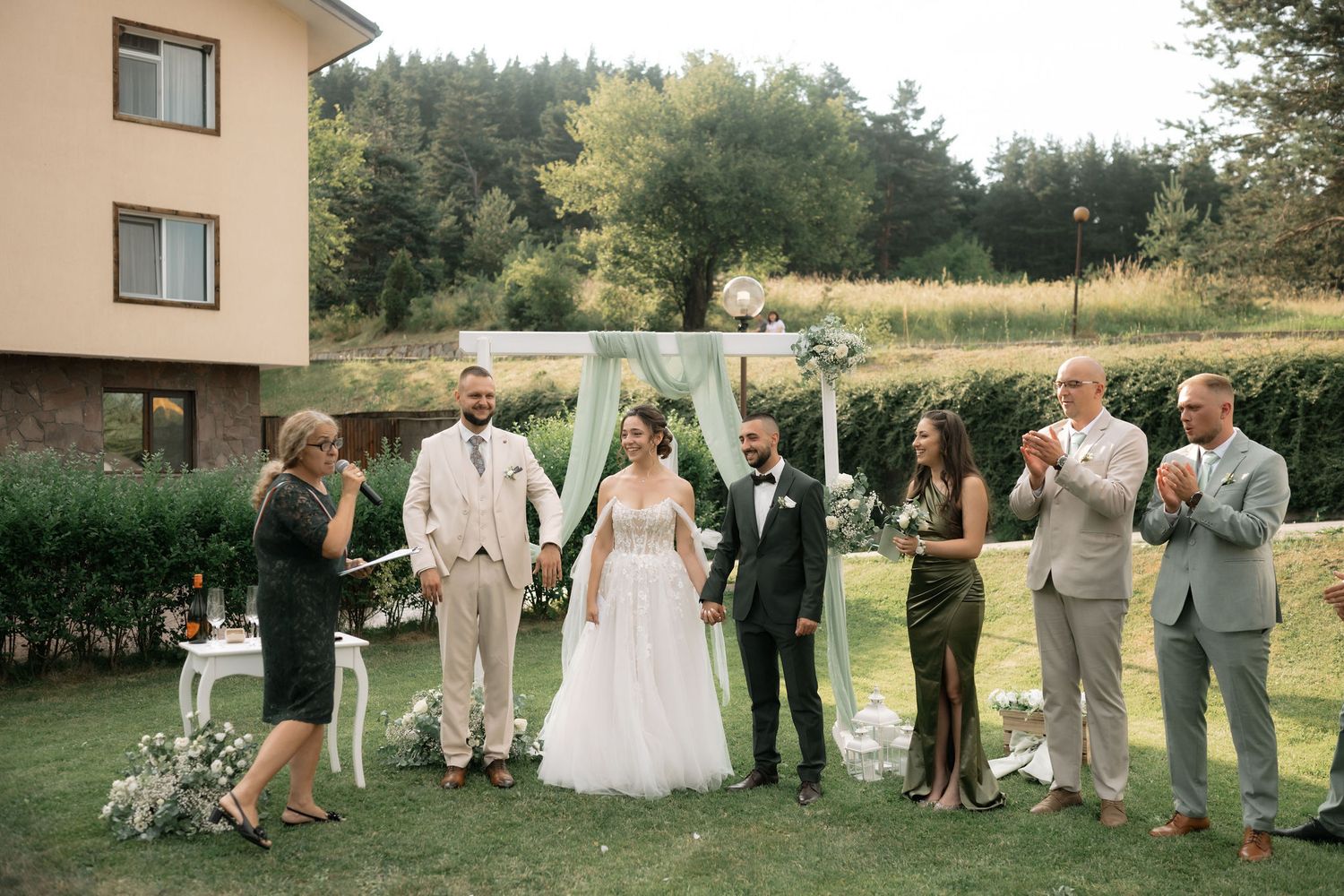 Wedding party in formal attire poses together on a grassy lawn outside during a summer celebration.