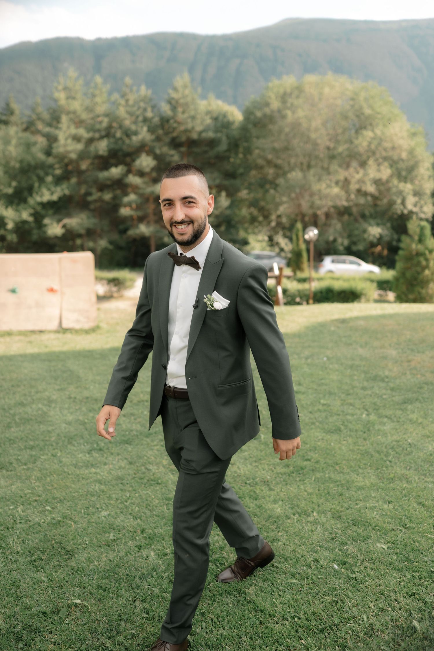 A person in a black tuxedo runs playfully across a grassy field with mountains in the background.