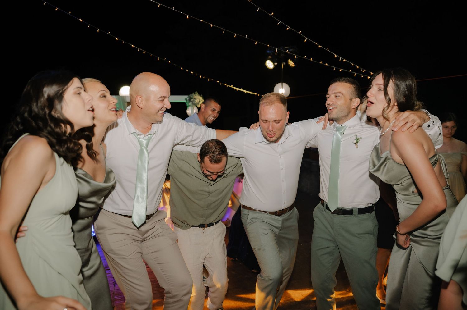 Wedding party guests dancing and laughing together in sage green attire at a reception celebration.