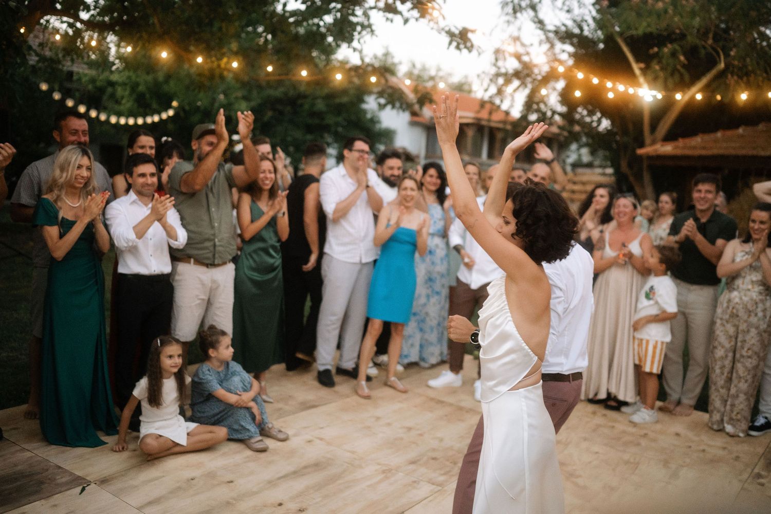 Wedding guests dance and celebrate at an outdoor reception with string lights at sunset.