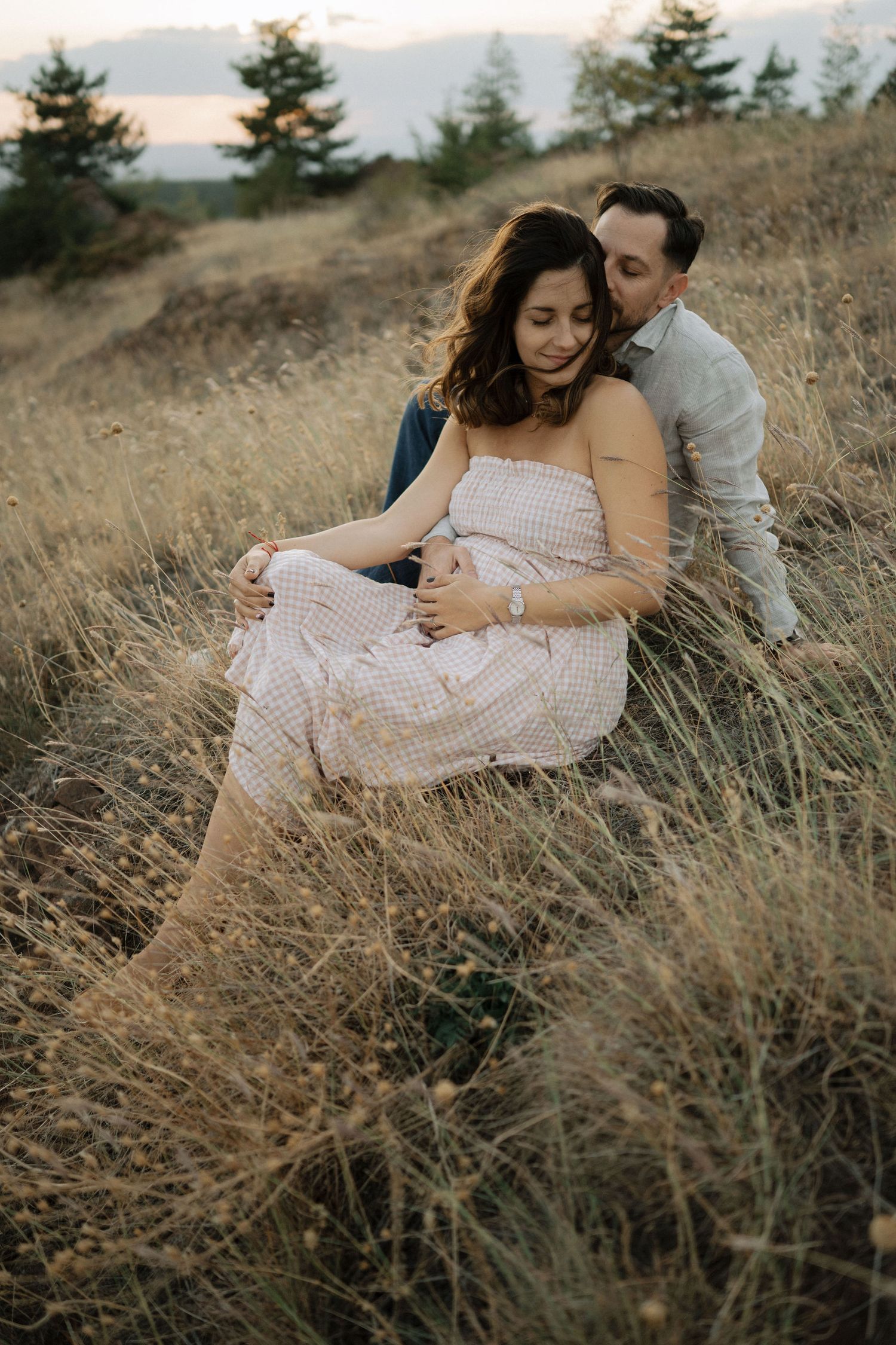 Couple sitting together in tall dried grass on a hillside during golden hour.
