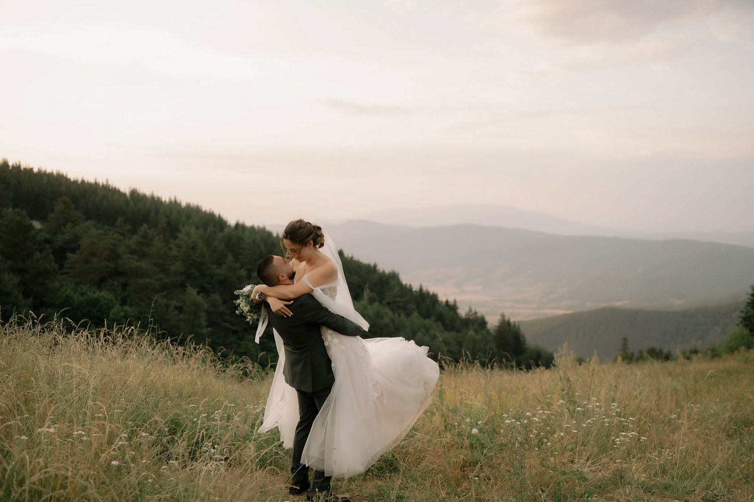 A couple in wedding attire dances together in a scenic mountain meadow at sunset.