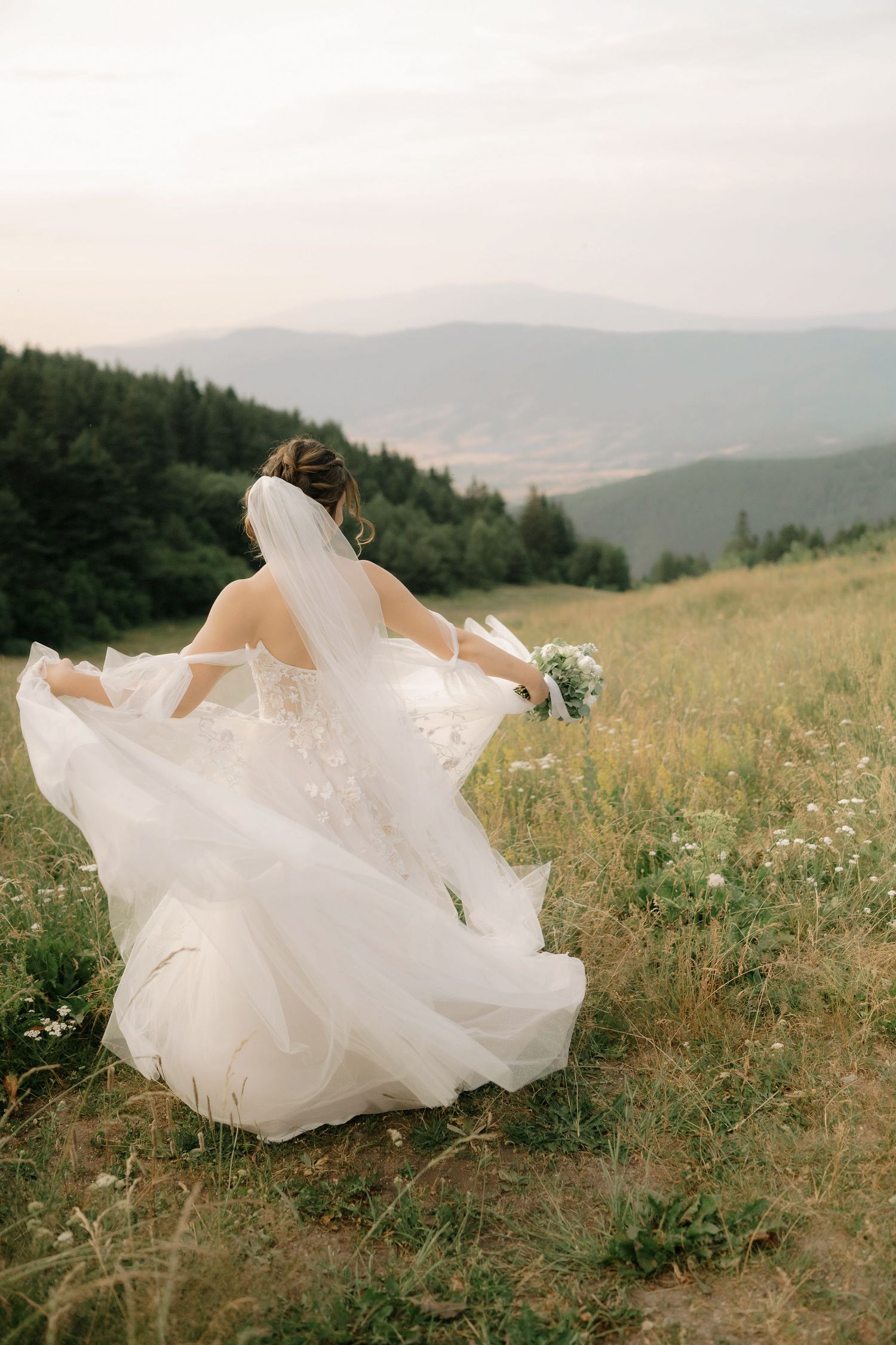 A flowing white wedding dress billows in the wind against a scenic mountain meadow landscape with wildflowers.