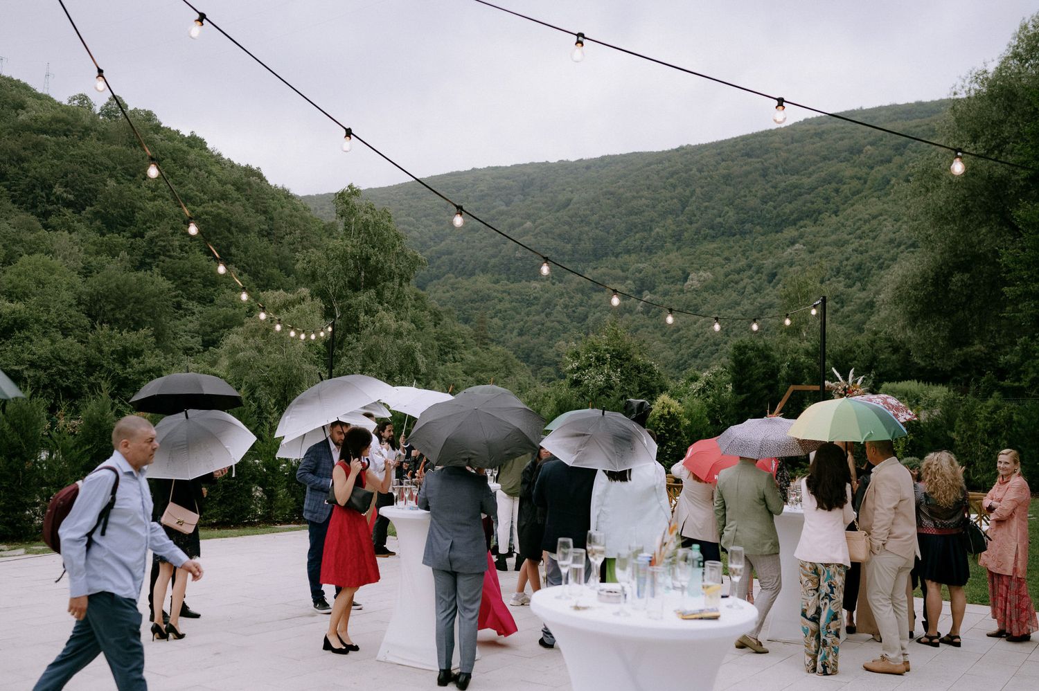 Wedding guests holding umbrellas gather outdoors on a rainy day in a lush mountain setting.