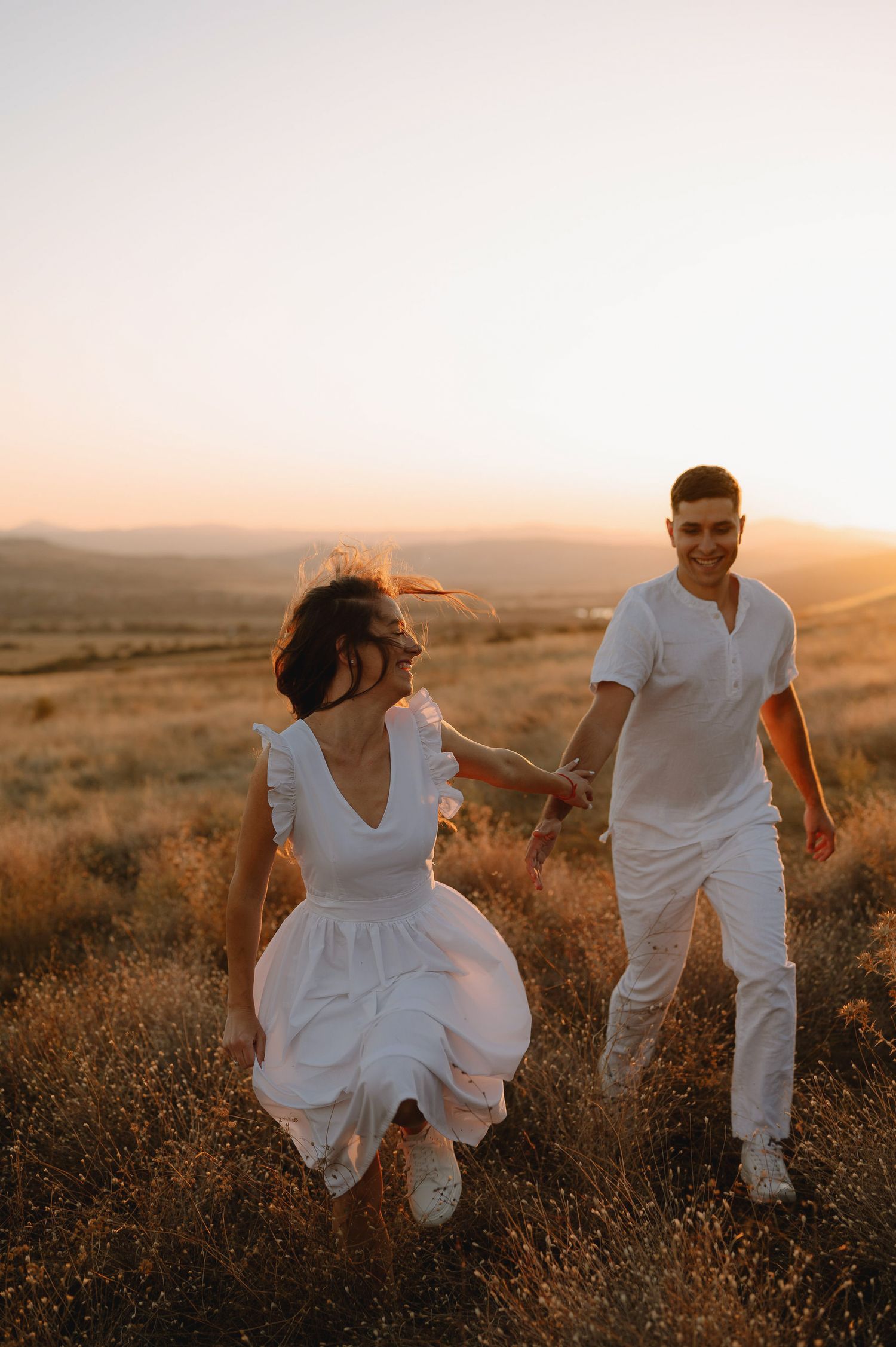 A couple in white outfits dances playfully together in a golden field at sunset.