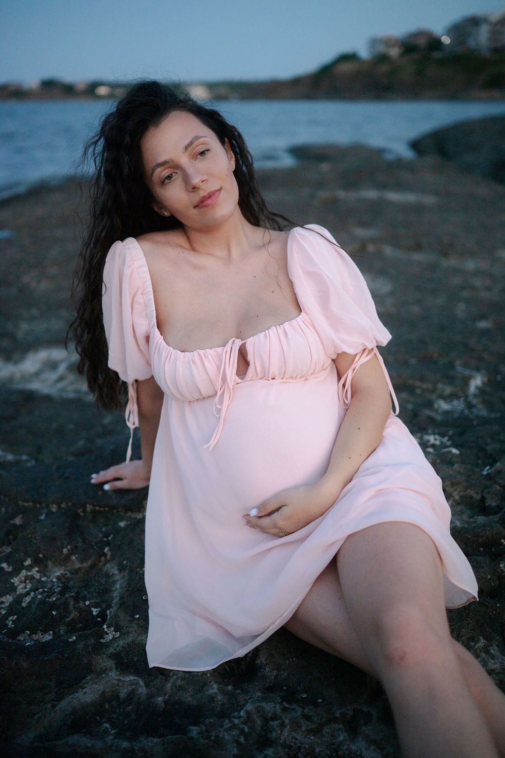 A pregnant woman in a pink dress sits on rocky coastal shoreline at sunset.