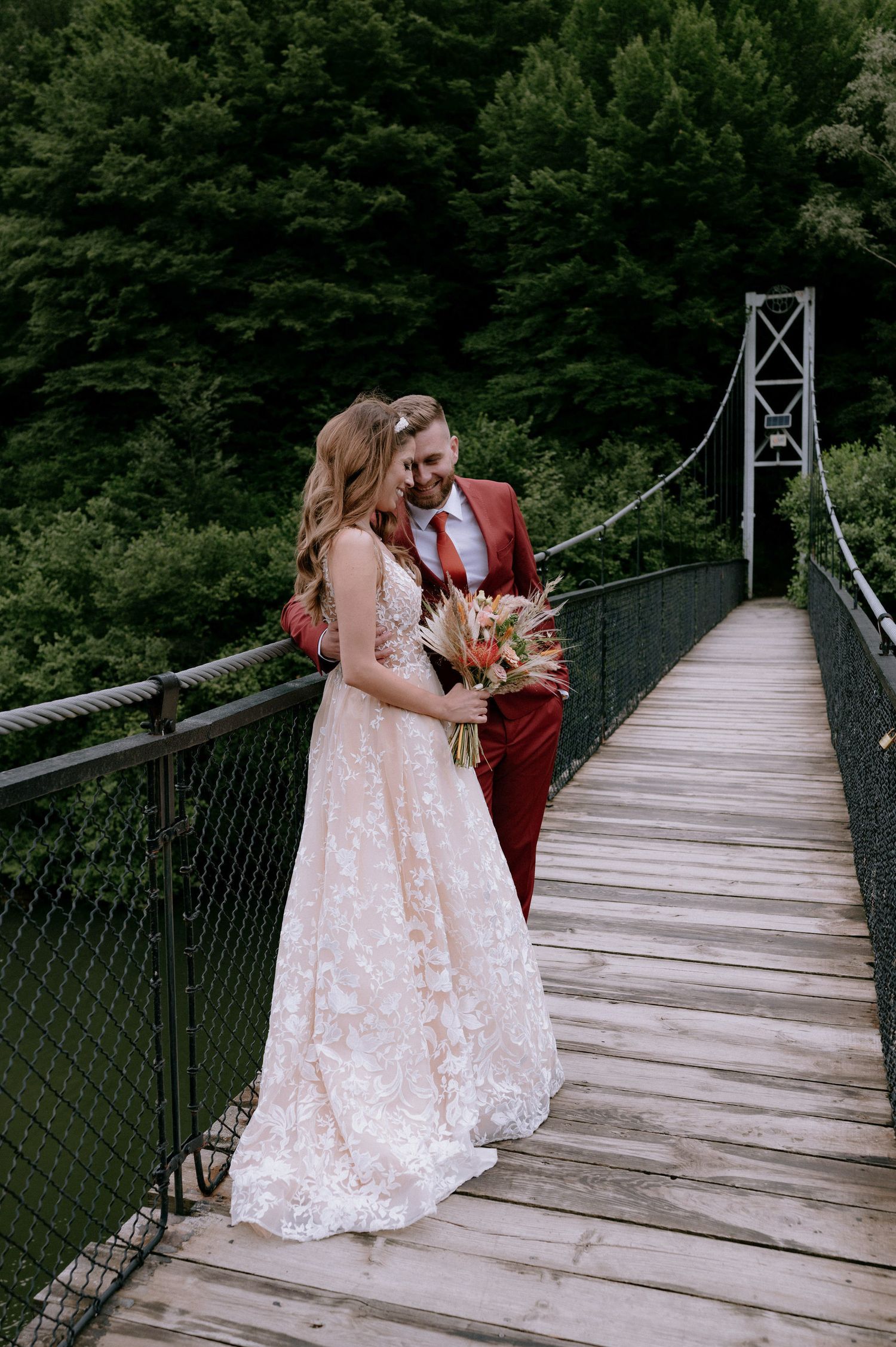 A couple in wedding attire stands together on a rustic wooden suspension bridge surrounded by lush green forest.