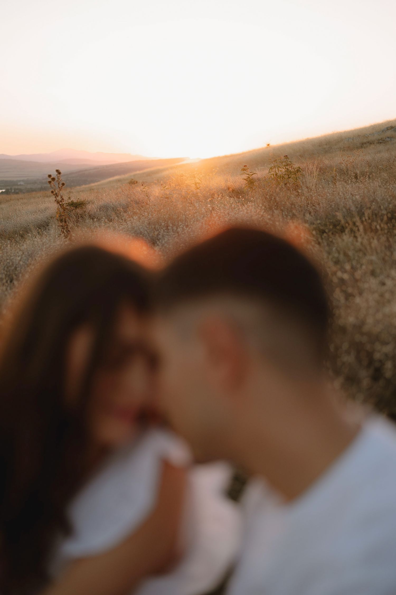 A couple embraces in silhouette against a golden sunset over rolling hills during a romantic outdoor photoshoot.