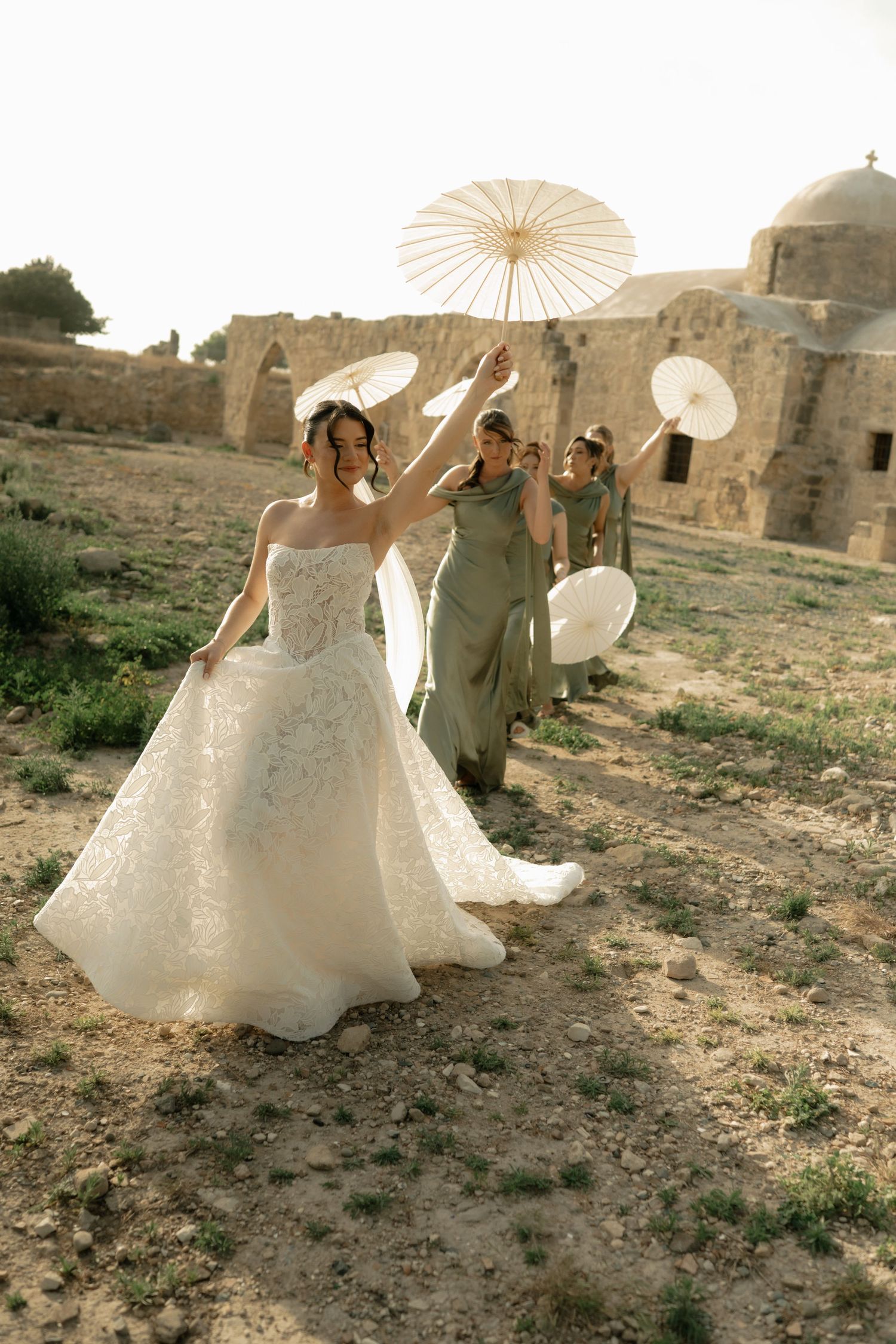 Bride in white lace gown walks with bridesmaids in sage green dresses holding parasols near ancient stone ruins.