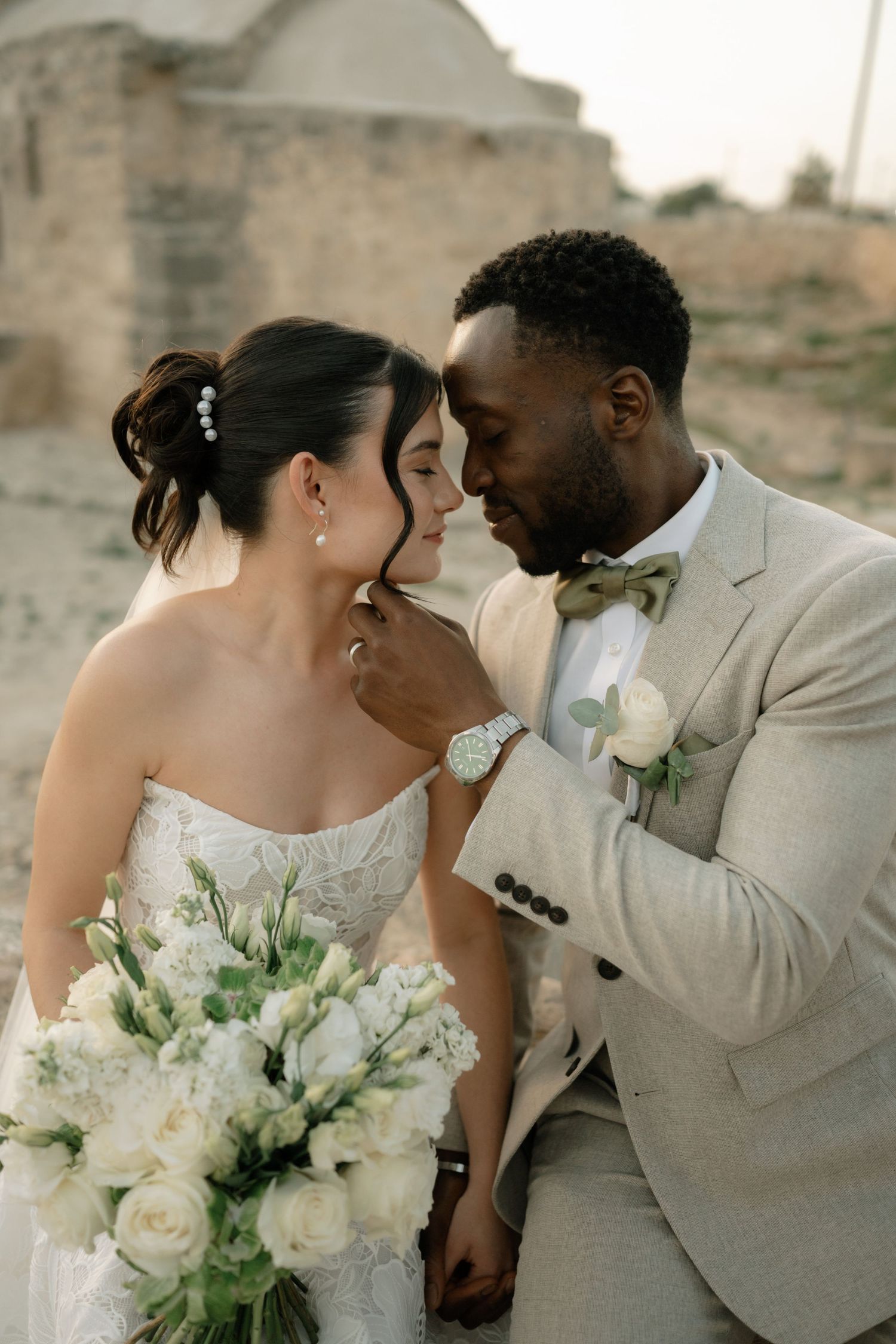 Interracial couple in wedding attire share romantic moment, groom in beige suit touches bride's face as she holds white bouquet.